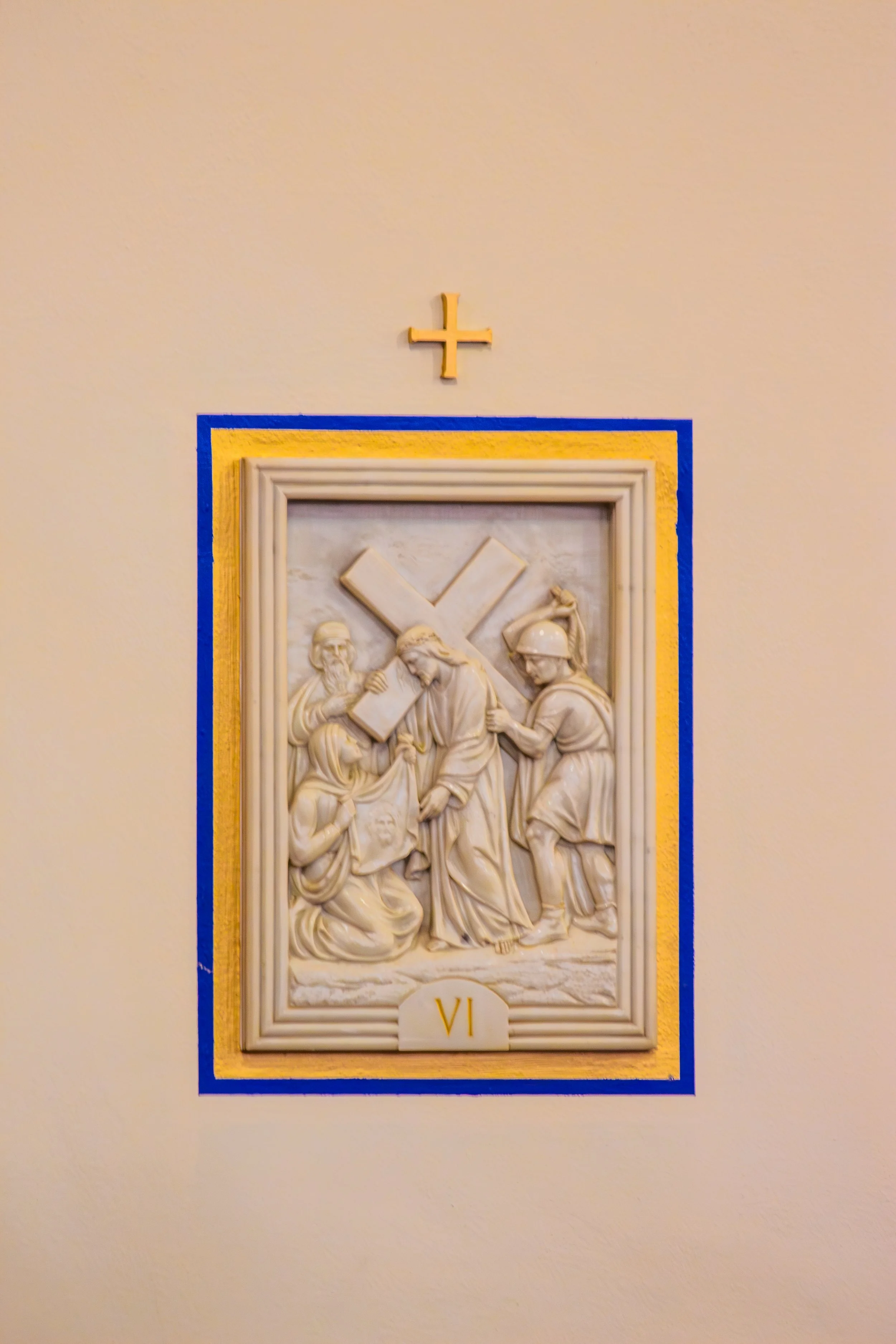 Relief sculpture depicting the biblical scene of Jesus carrying the cross, with Roman soldiers and followers, framed with a yellow and blue border, and a small cross above.