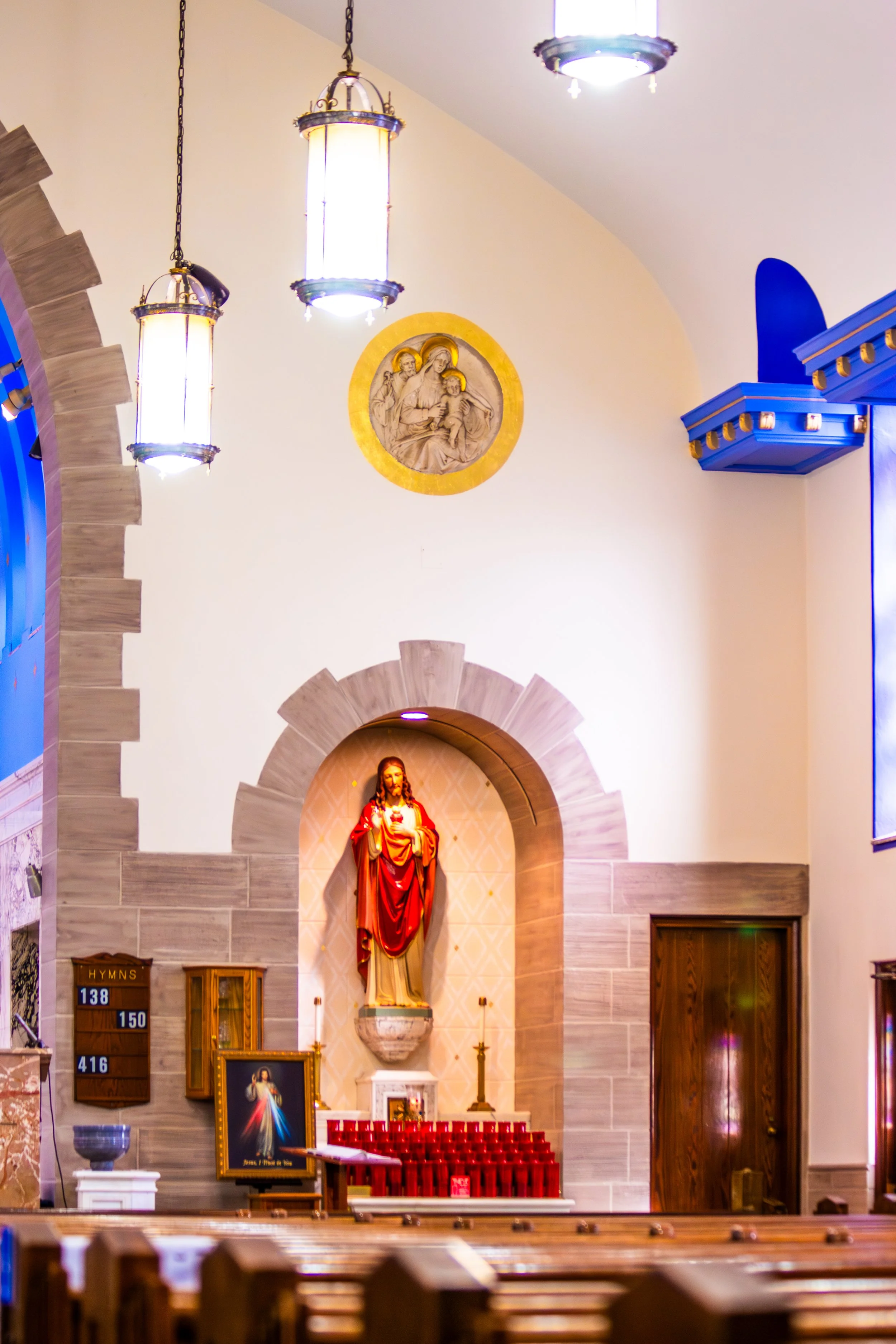 Interior of a church with a prominent statue of Jesus and several religious symbols and icons. There are hanging lights and colorful decorations, along with hymn boards on the left side.