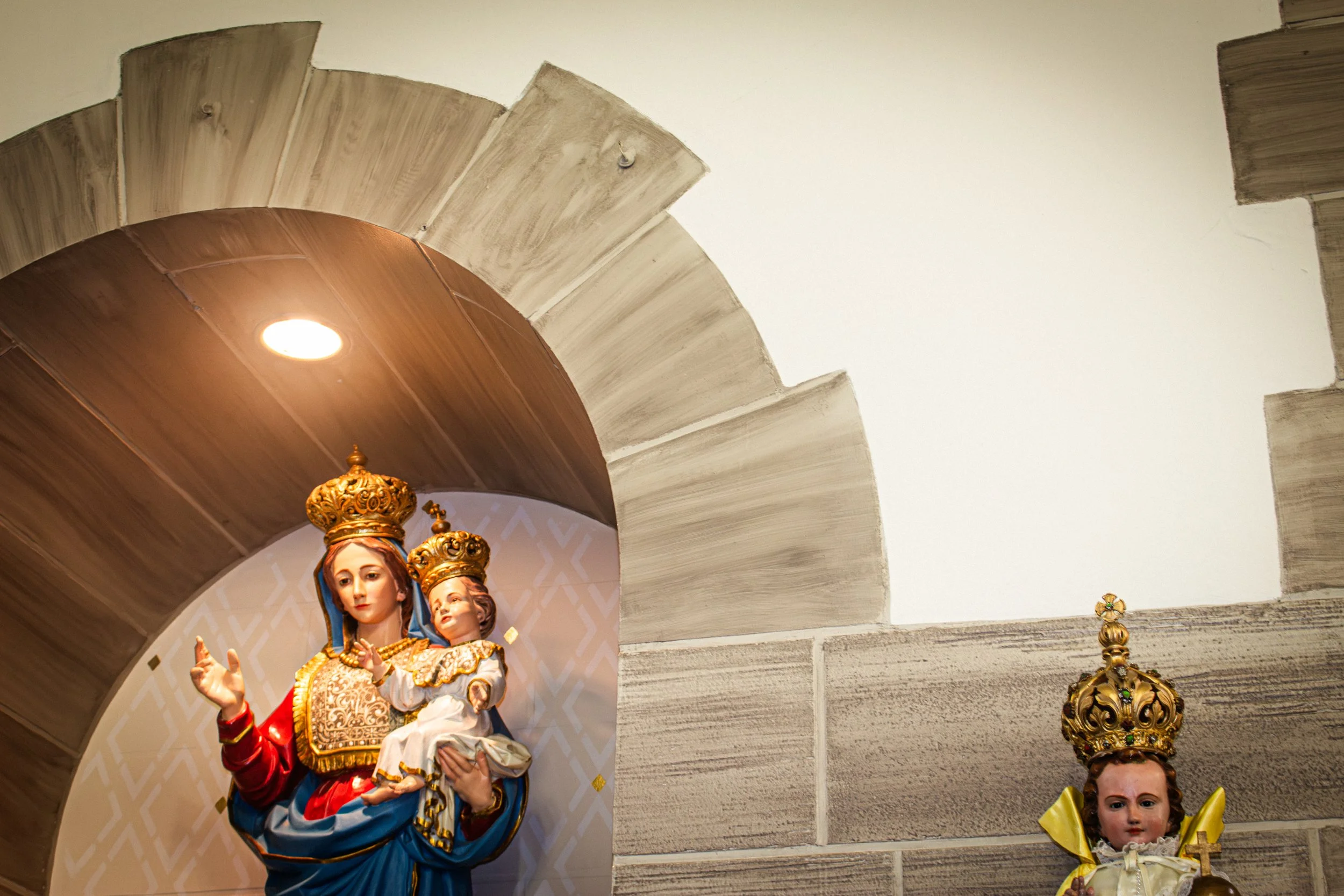 Religious statues, including Virgin Mary holding baby Jesus, with ornate gold crowns and colorful clothing, placed on a ledge under a wooden and stone arch ceiling.