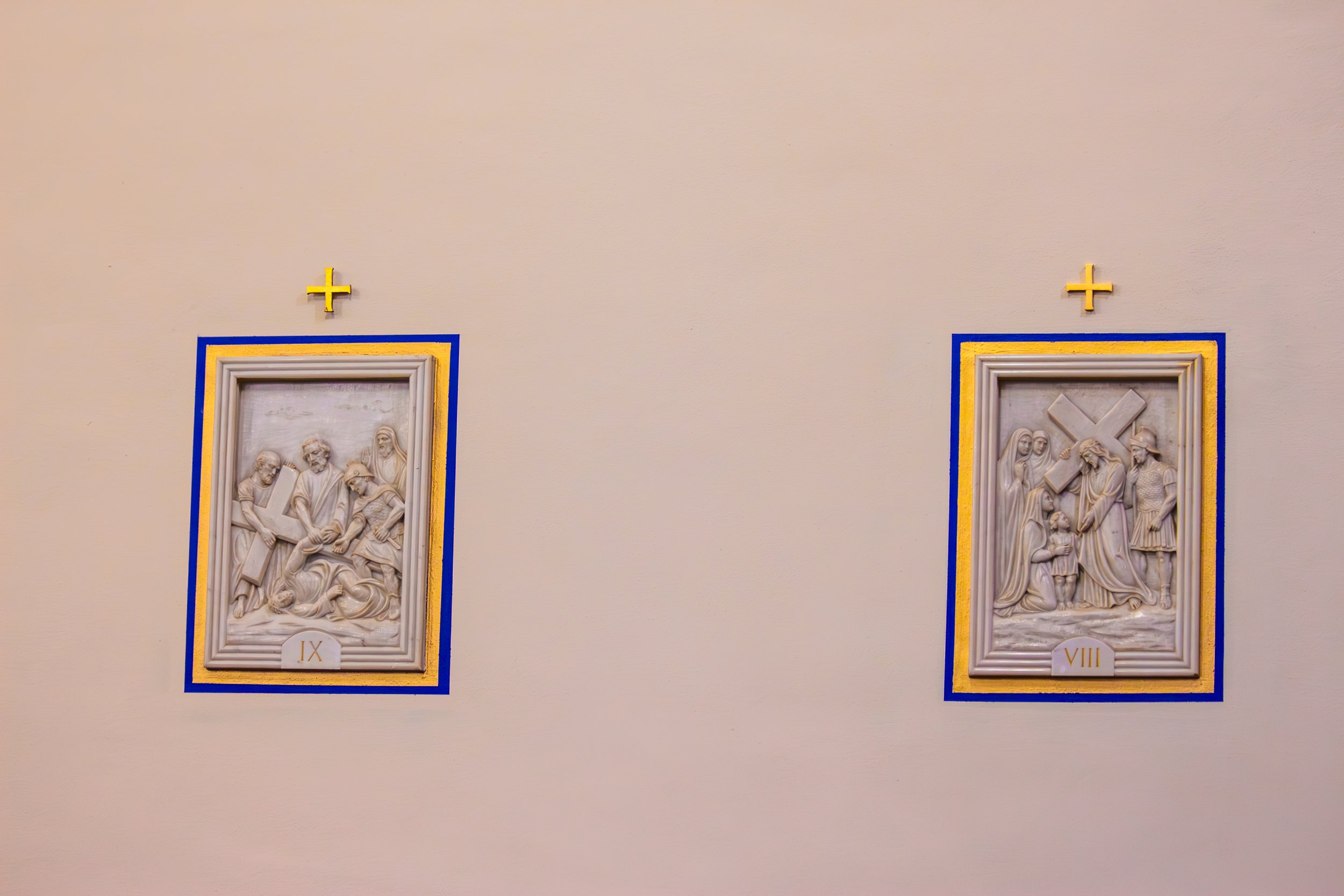 Two religious relief sculptures on a plain wall, framed with blue and yellow borders, each with Roman numerals IX and VIII, and adorned with small gold crosses above them.