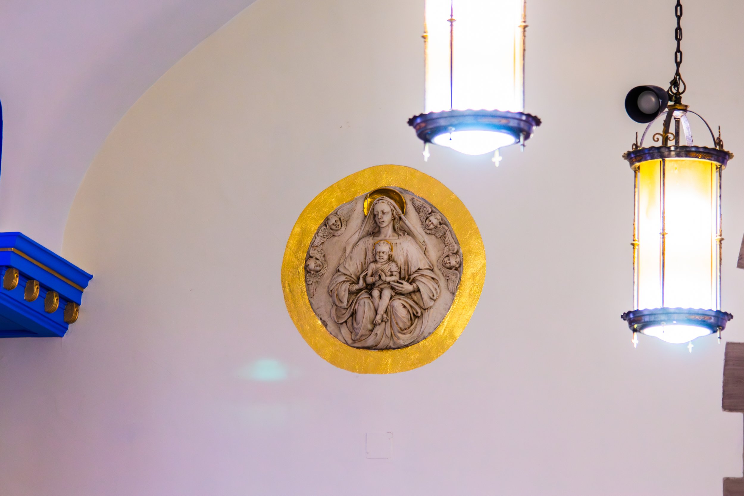 Religious wall sculpture of the Virgin Mary holding baby Jesus, surrounded by angels with a gold background, along with hanging pendant lamps illuminating the space.
