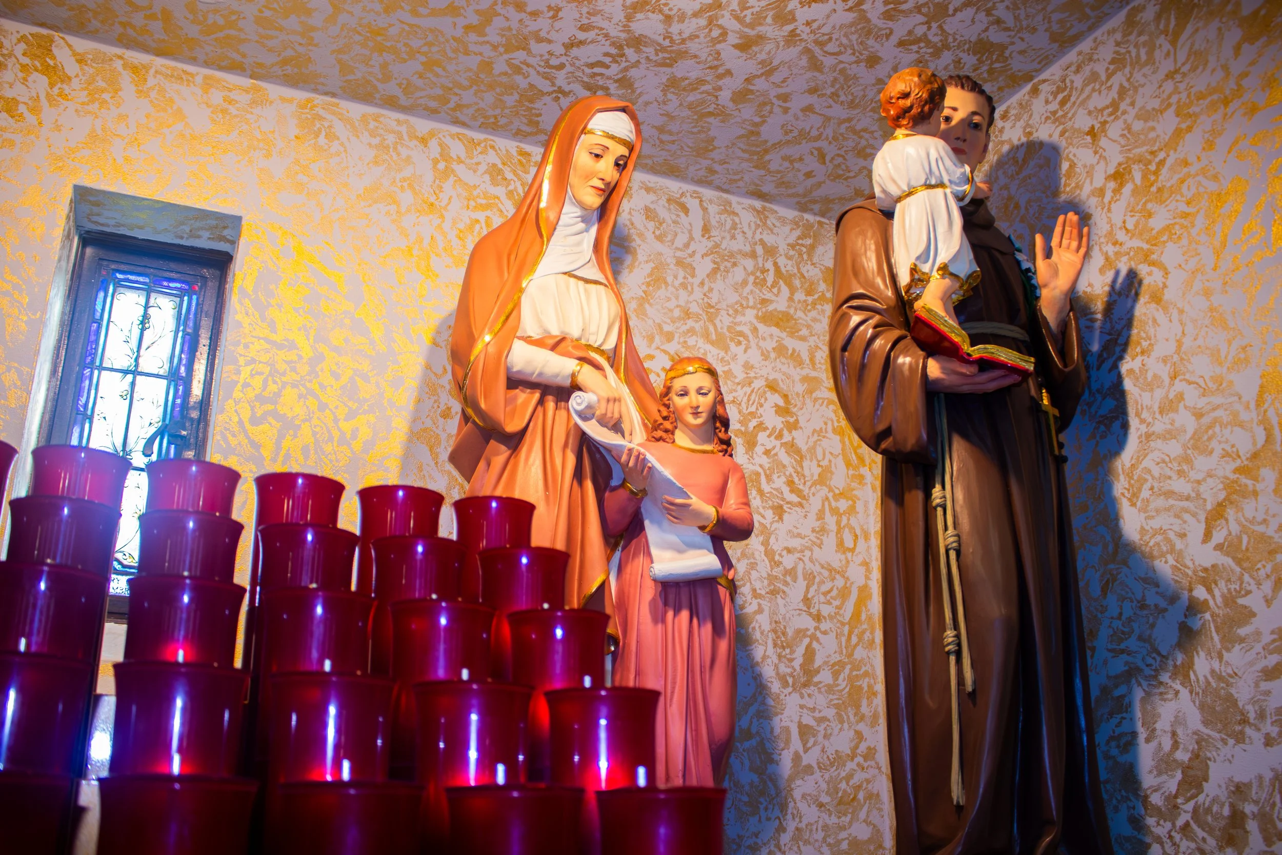 Religious statues on a wall with a window and red candles in front.