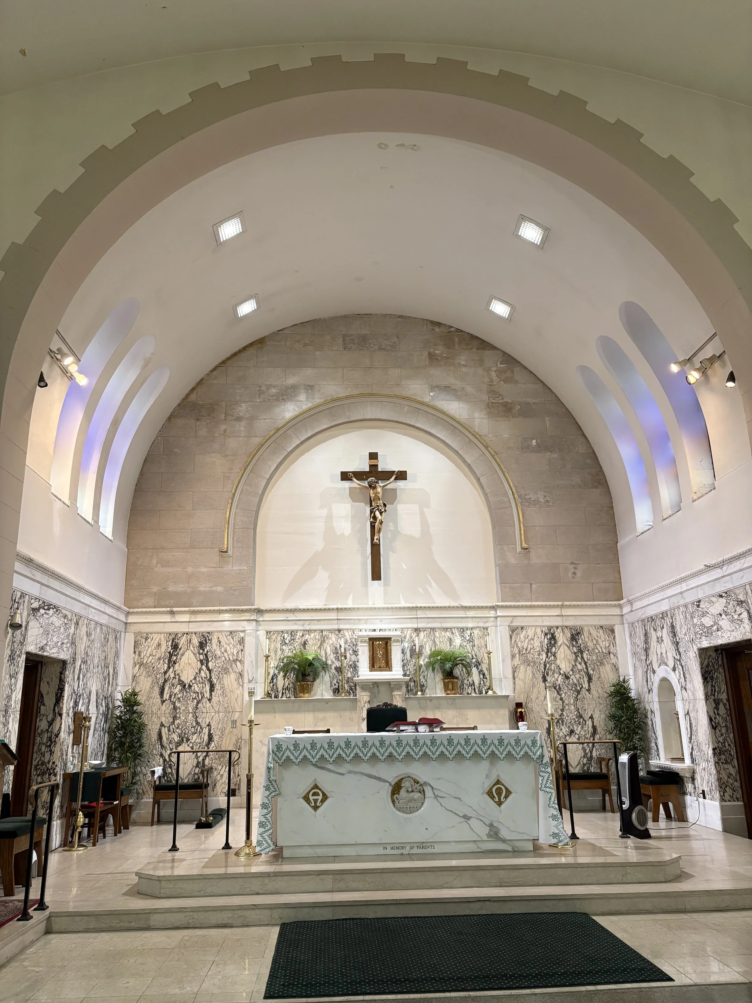 The interior of a church or chapel with an altar, crucifix, and religious symbols, decorated with marble and plants.