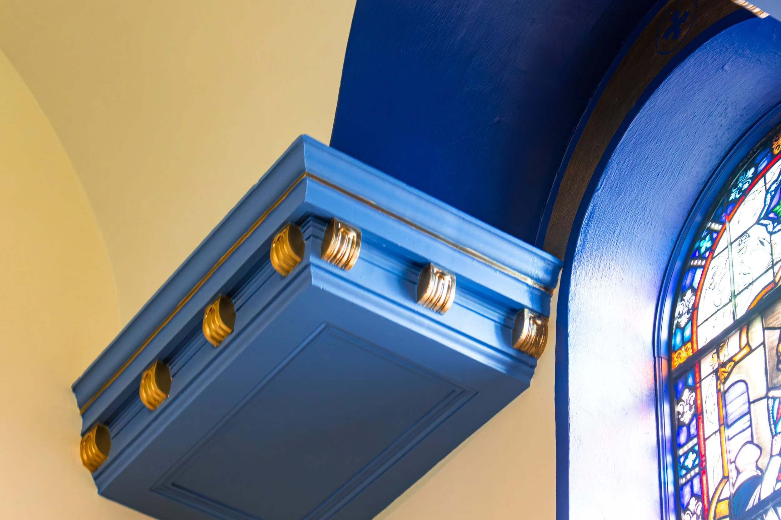 Close-up of a blue bookshelf with gold accents next to a stained glass window with colorful designs.