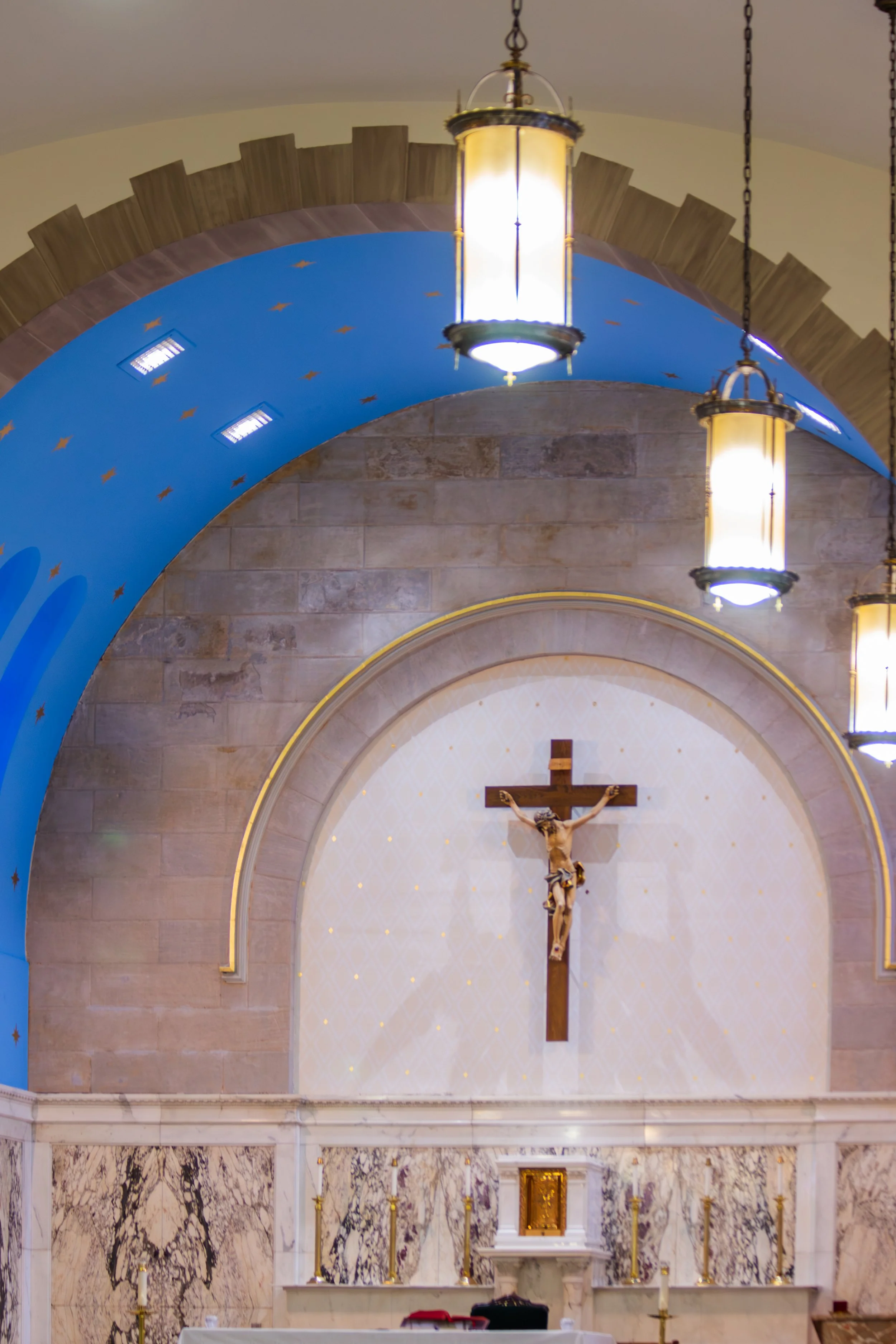 Interior view of a church altar with a crucifix and lamps, featuring a blue and gold arch with star decorations and marble walls.