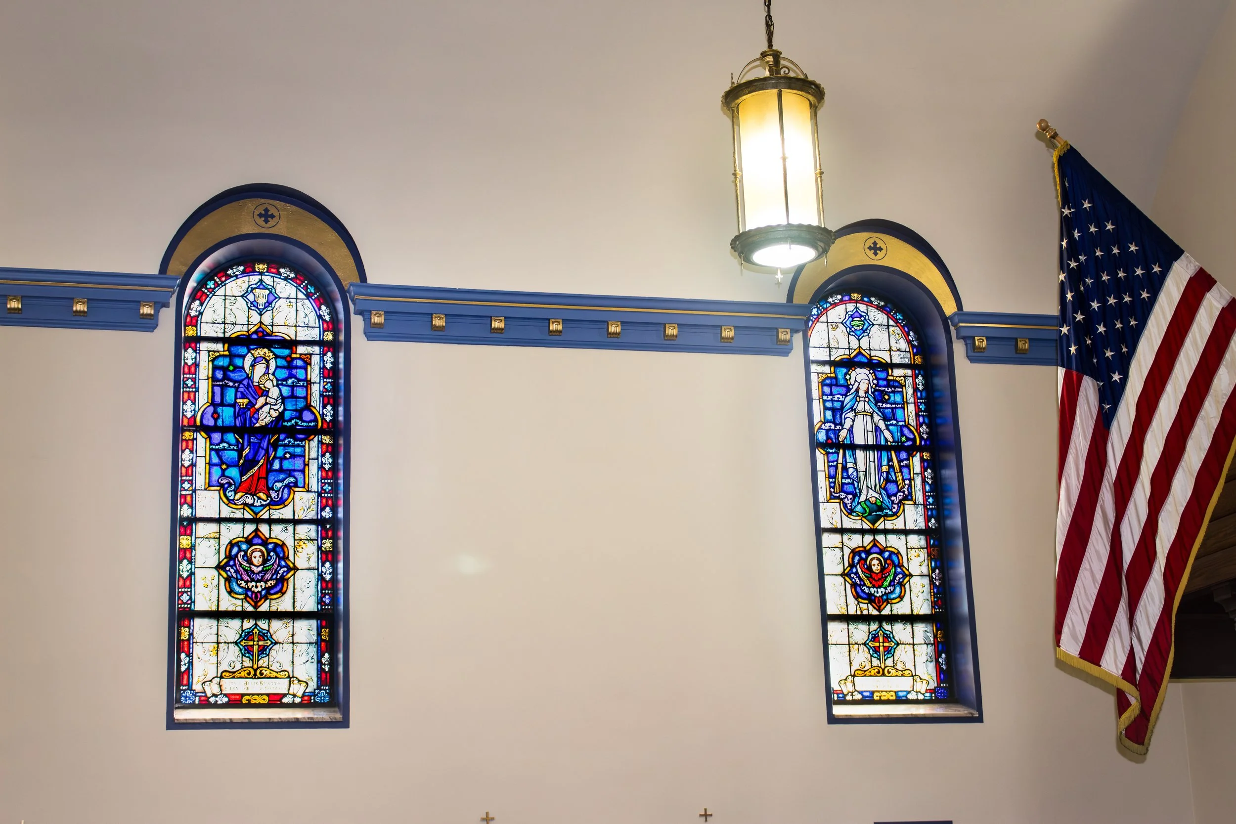 Interior of a church with two stained glass windows depicting religious figures, a hanging lantern-style light, an American flag on the right, and small crosses on the wall