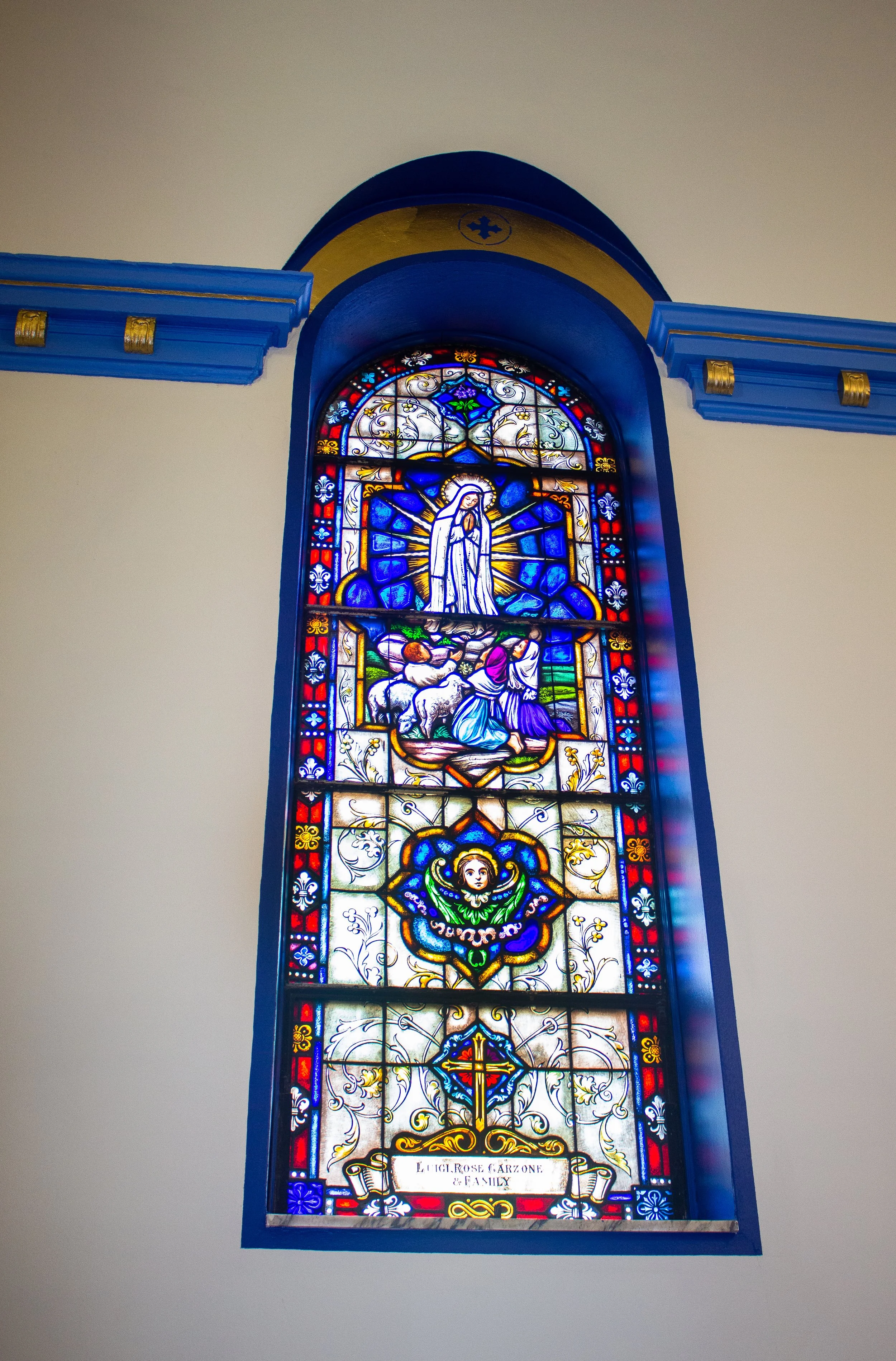 Colorful stained glass window with religious imagery and decorative patterns, framed in blue, with a title at the bottom reading "Lucie Rose Faizone & Family."