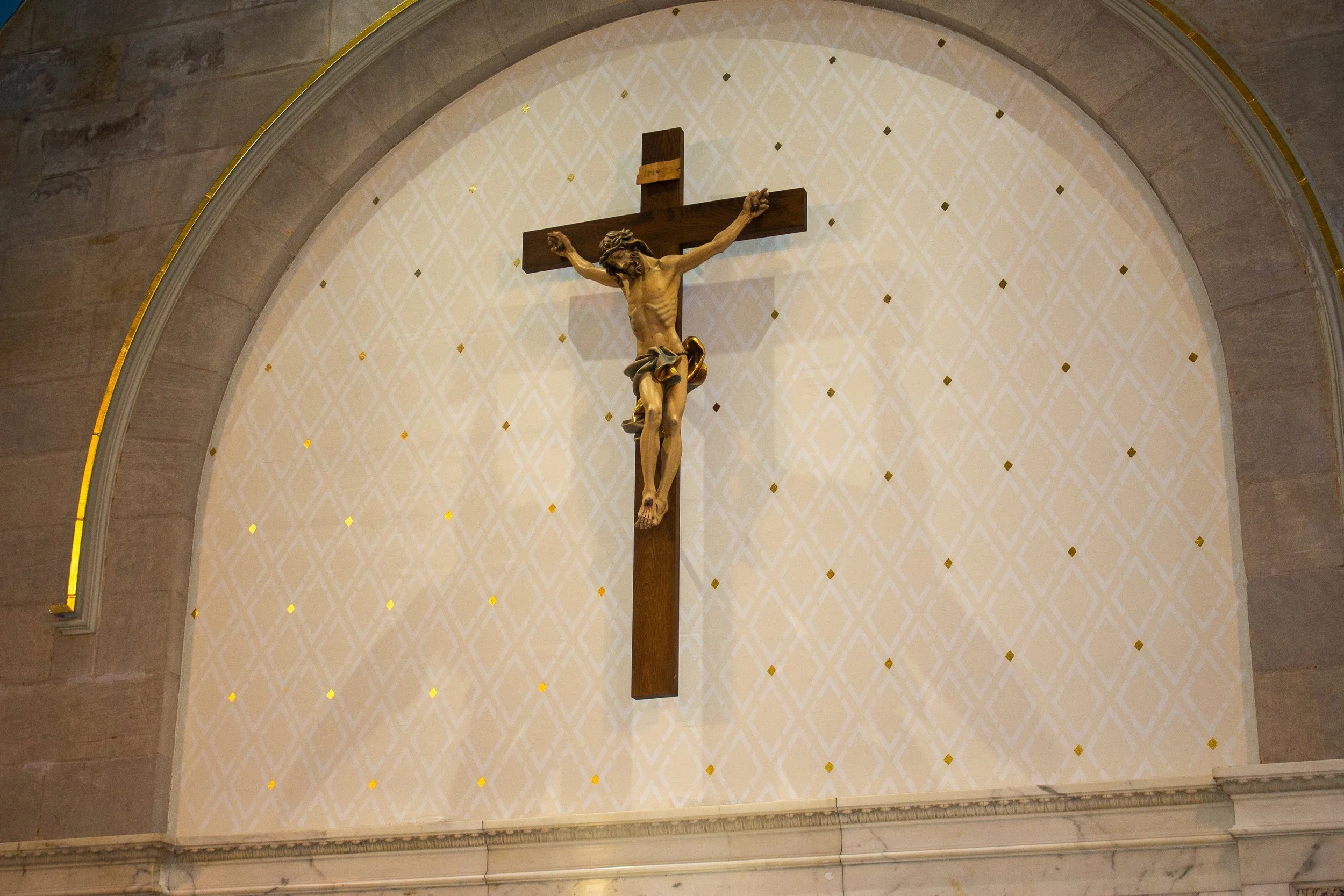 A crucifix with Jesus Christ on the cross mounted on a wall inside a church or cathedral, above an arch with a patterned background.