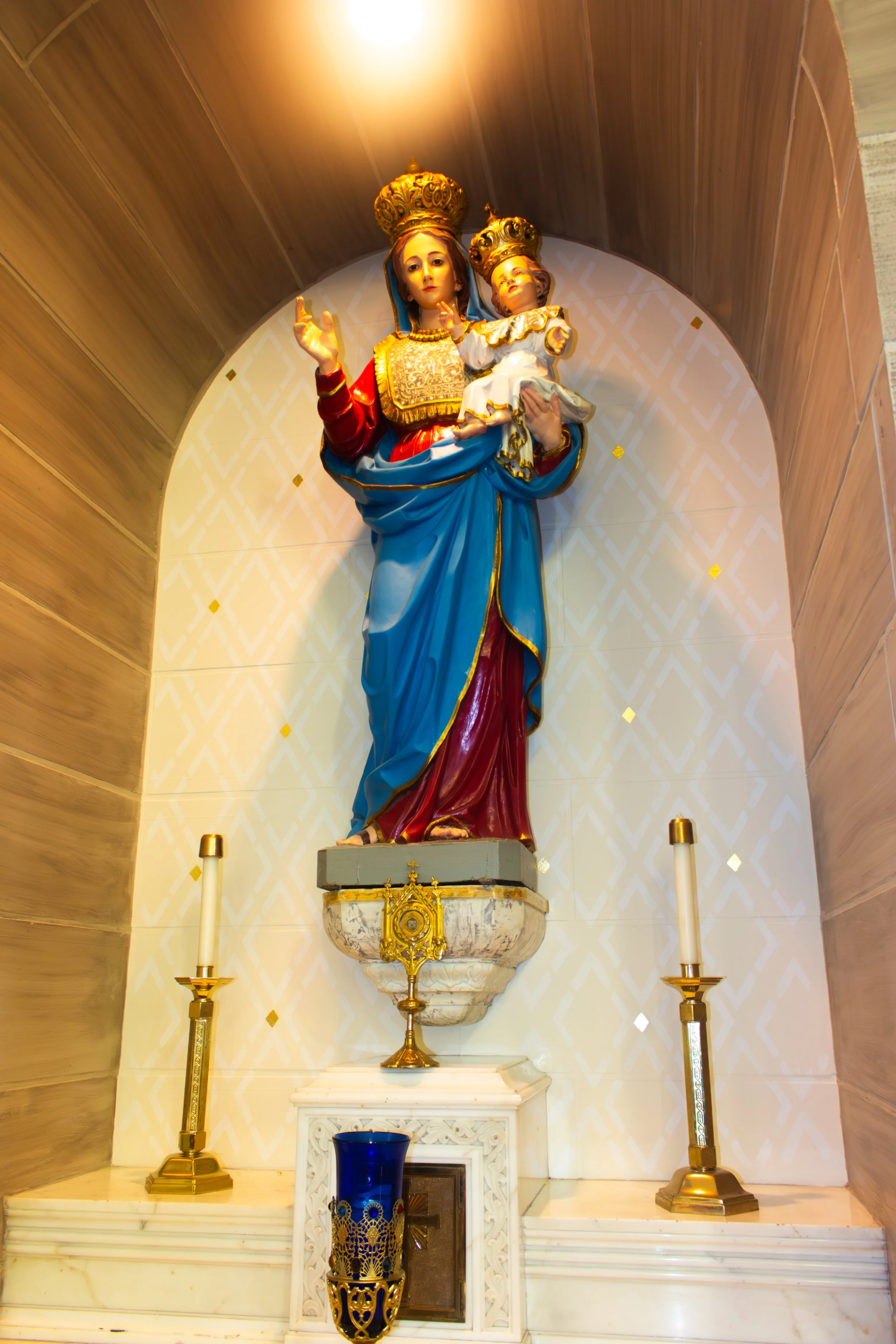 Statue of the Virgin Mary holding baby Jesus, decorated with gold crowns and garments, flanked by two gold candlesticks, placed on a marble altar with a blue glass candle holder in front.