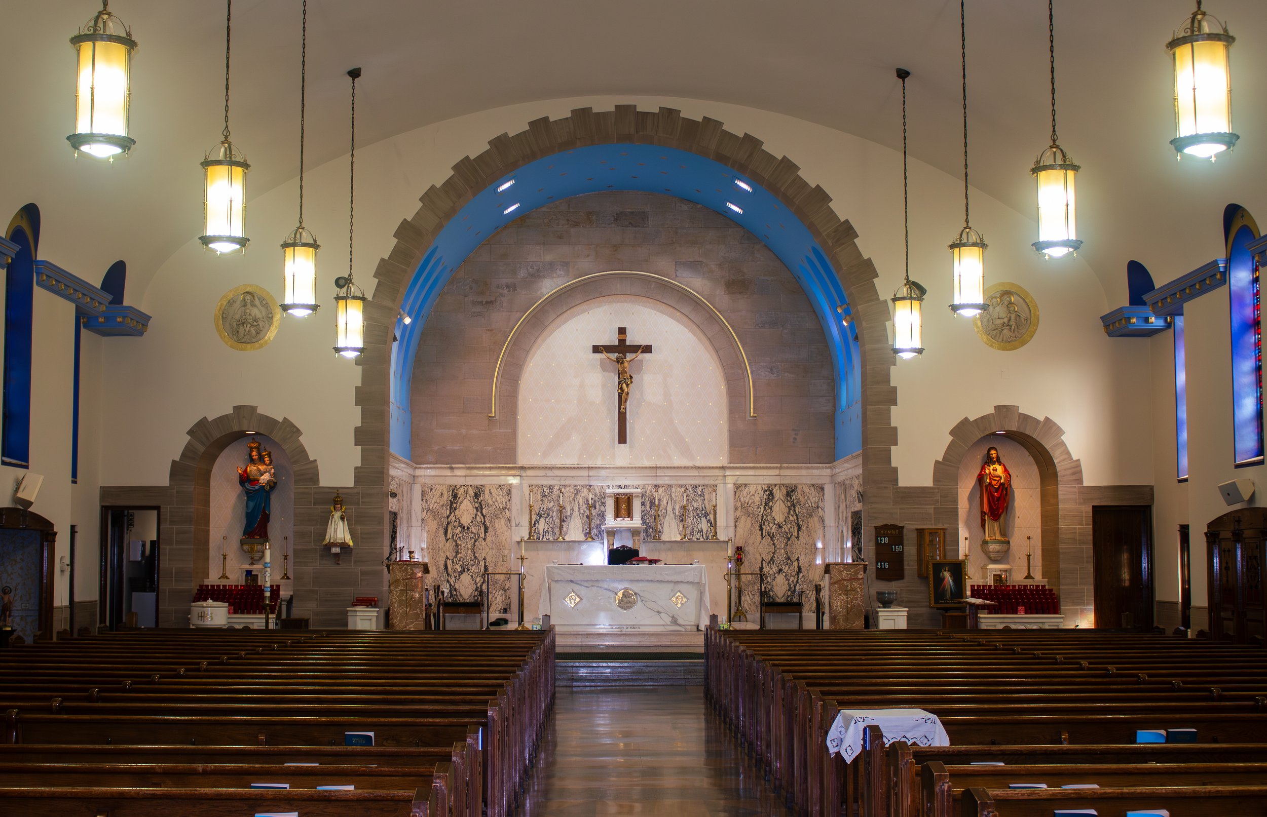 The interior of a church with wooden pews, an altar with religious statues, a crucifix, and religious artwork on the walls.