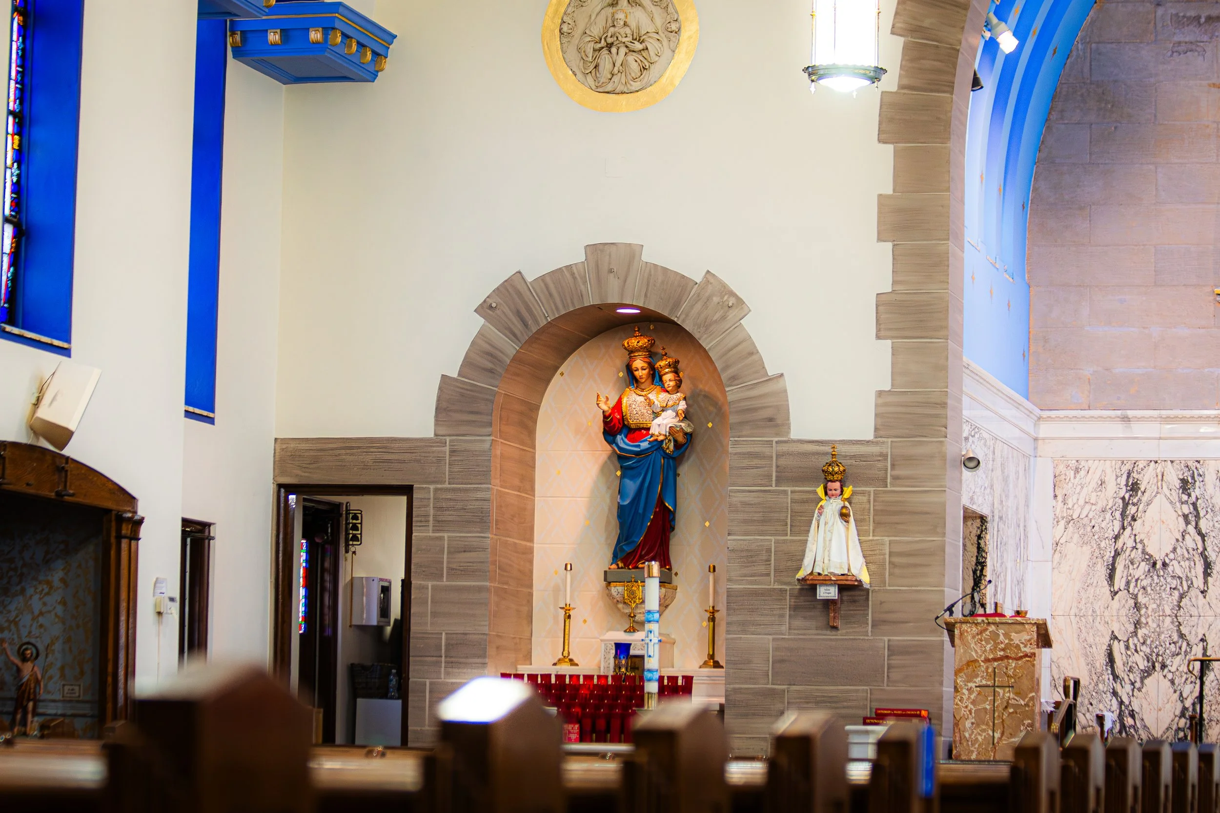 Inside a church with religious statues of Mary and Jesus behind the altar, featuring a carved stone archway, candles, and colorful stained glass windows.