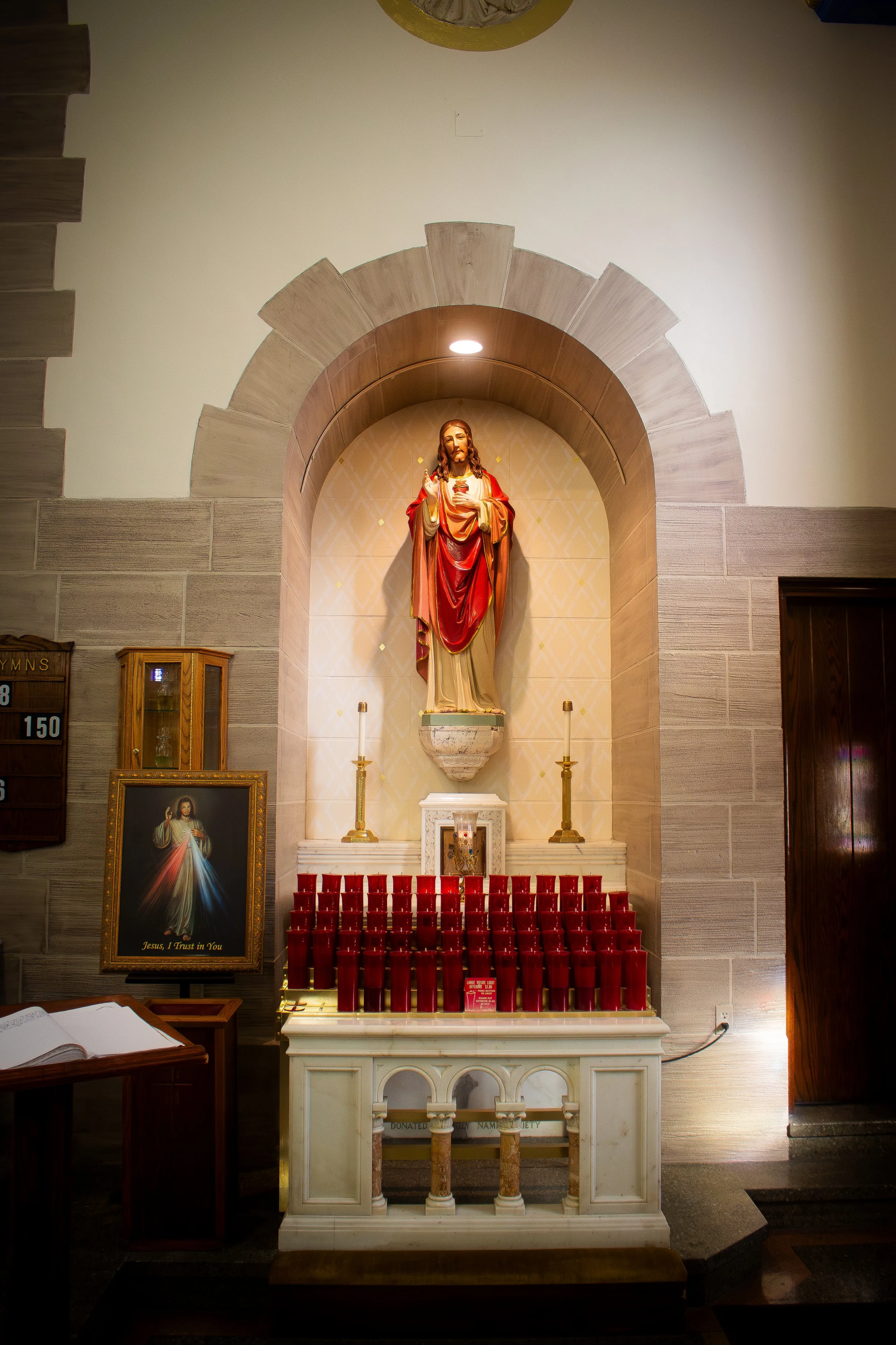 Inside a church or chapel, a statue of Jesus Christ holding a sacred heart and a finger raised in blessing, placed in a niche with a semi-circular arch. In front of the statue, an altar with multiple red votive candles. To the left, a framed picture 