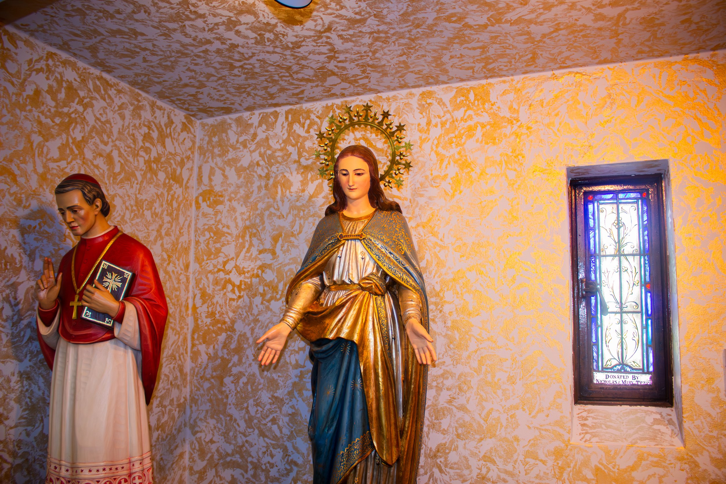 Three religious statues inside a church, including one of the Virgin Mary with a halo, another of a man holding a book, and a stained glass window on the right.