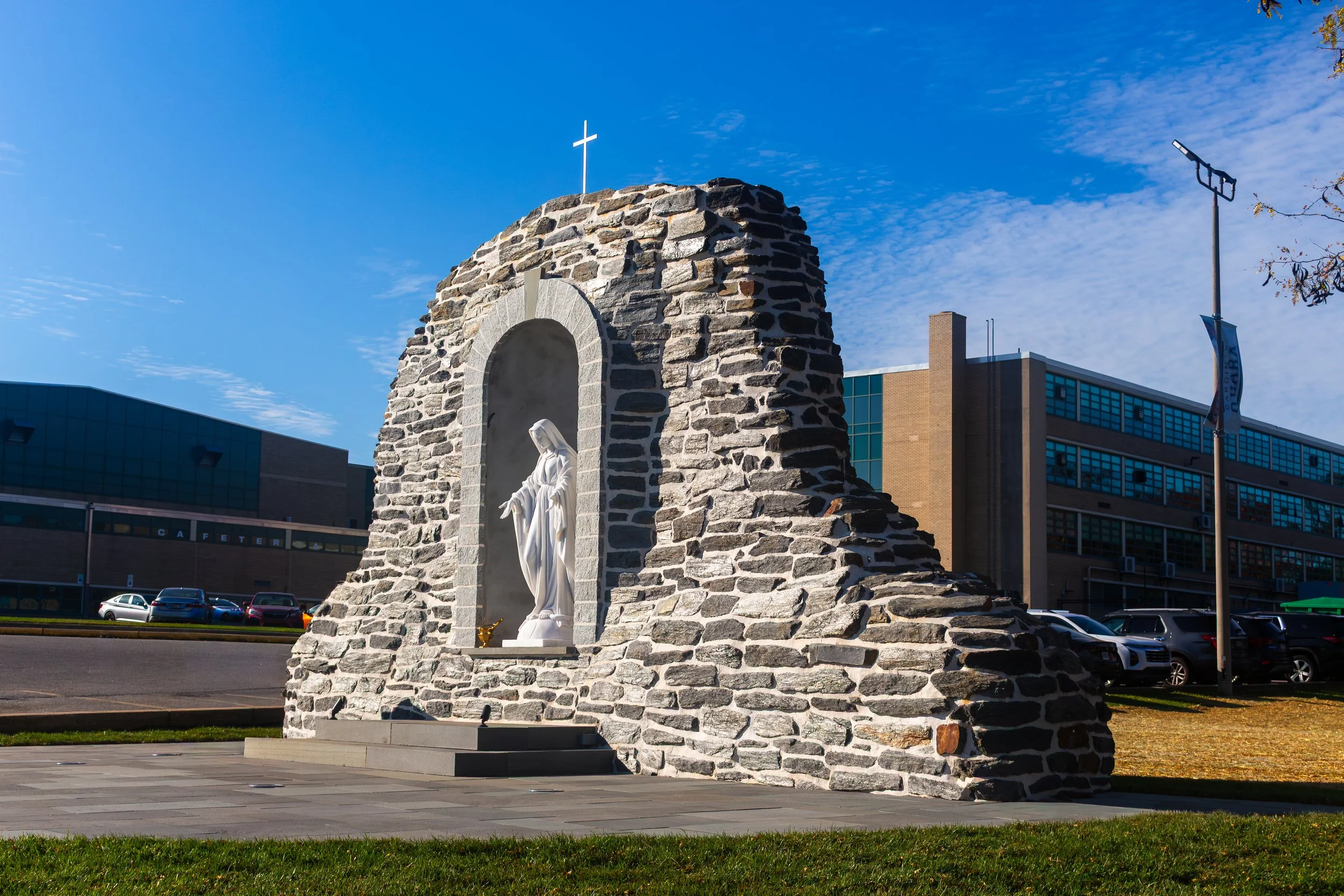 A stone shrine with statues of the Virgin Mary and a kneeling figure, surrounded by flowers and memorial plaques at the base, under a clear blue sky.