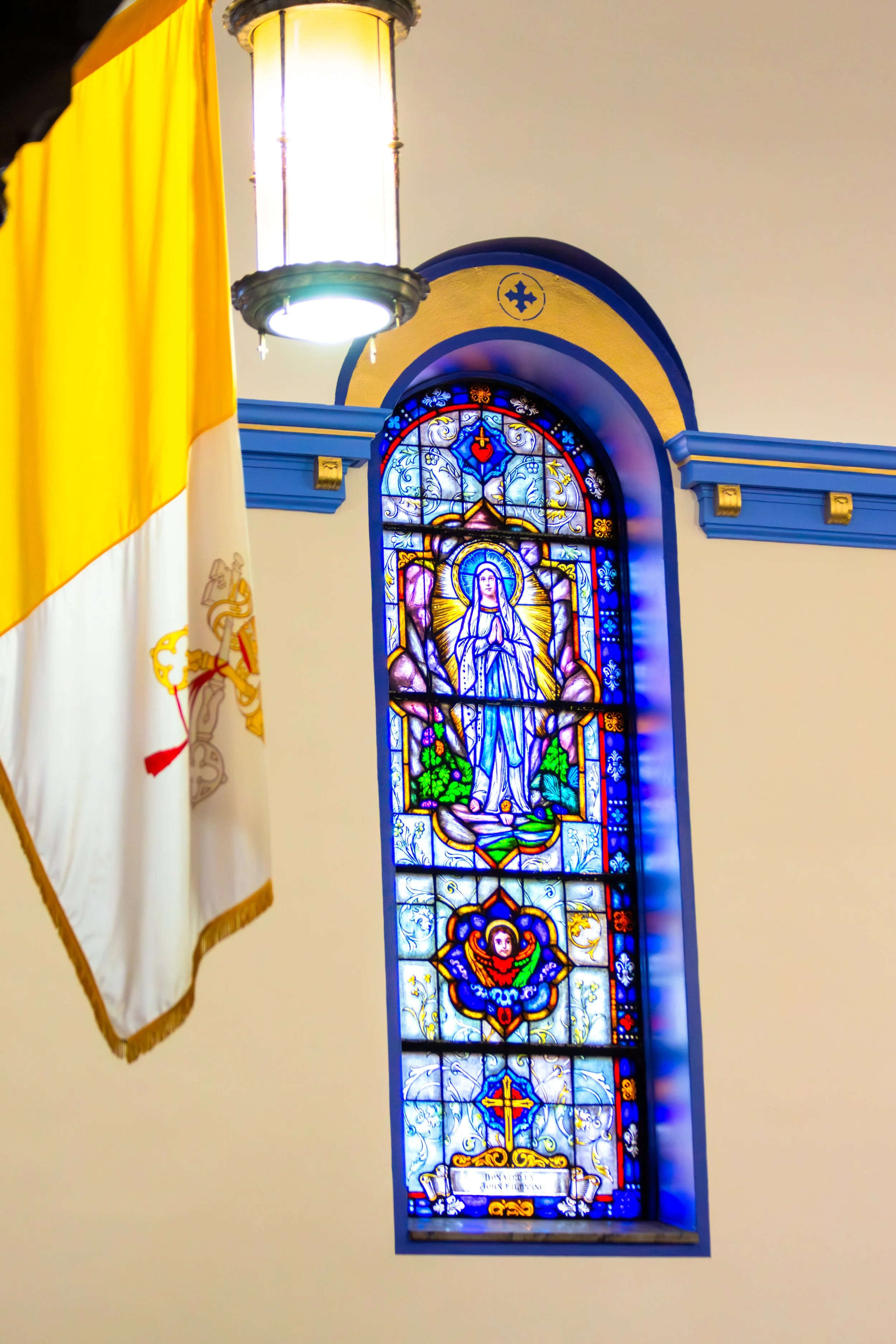 Close-up of a stained glass window depicting a religious figure, likely the Virgin Mary, with vibrant blue, gold, and white colors, surrounded by intricate designs and symbols. Part of a yellow and white flag and a hanging lantern are also visible in