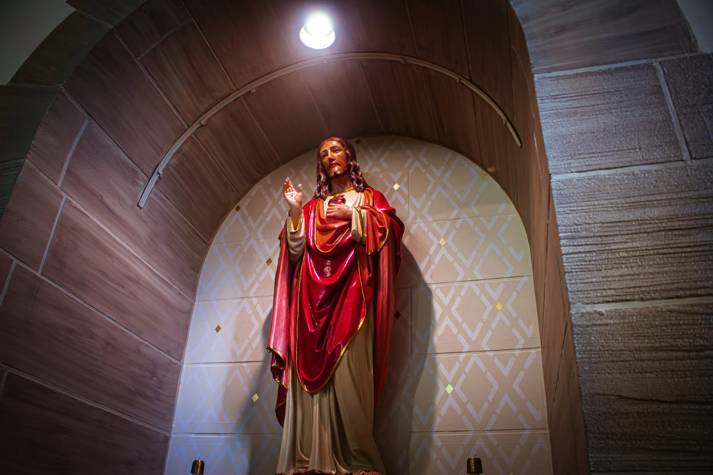 Statue of Jesus Christ with one hand raised and holding his heart, placed in a small chapel with wood-paneled walls and a decorative background.