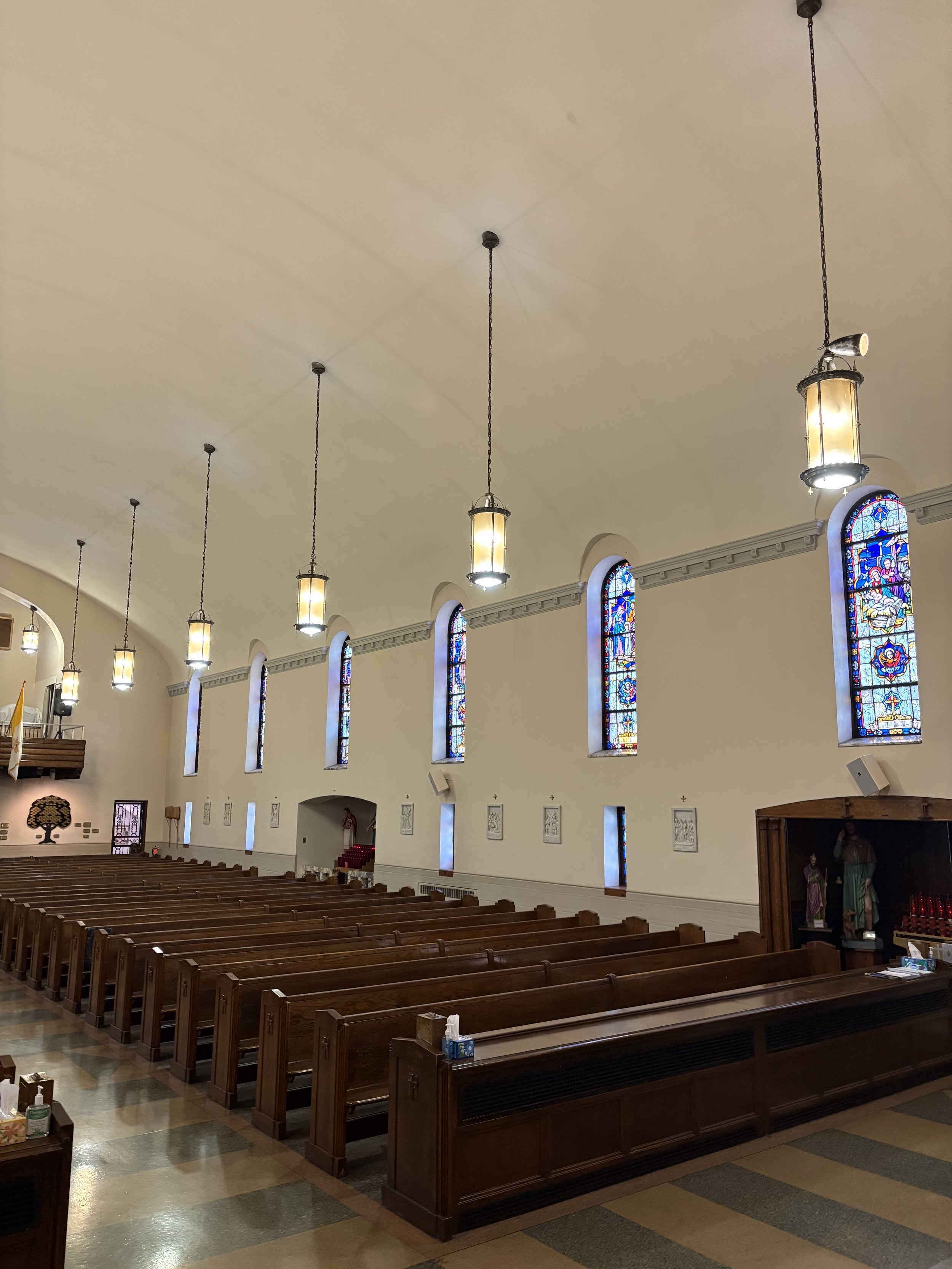 Inside a church, empty pews are lined up facing an altar, with stained glass windows along the wall and hanging lantern lights from the ceiling.