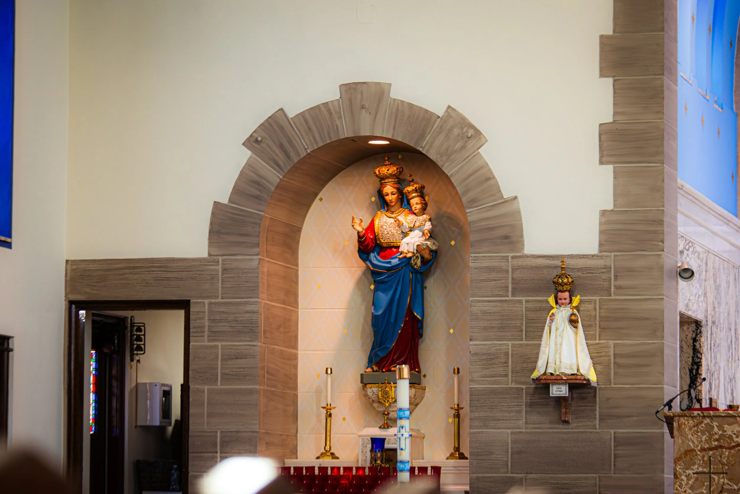 A religious altar featuring a statue of the Virgin Mary holding baby Jesus, both adorned with crowns. The altar is decorated with candles, gold accents, and a backdrop of arched wood and patterned wallpaper inside a church.