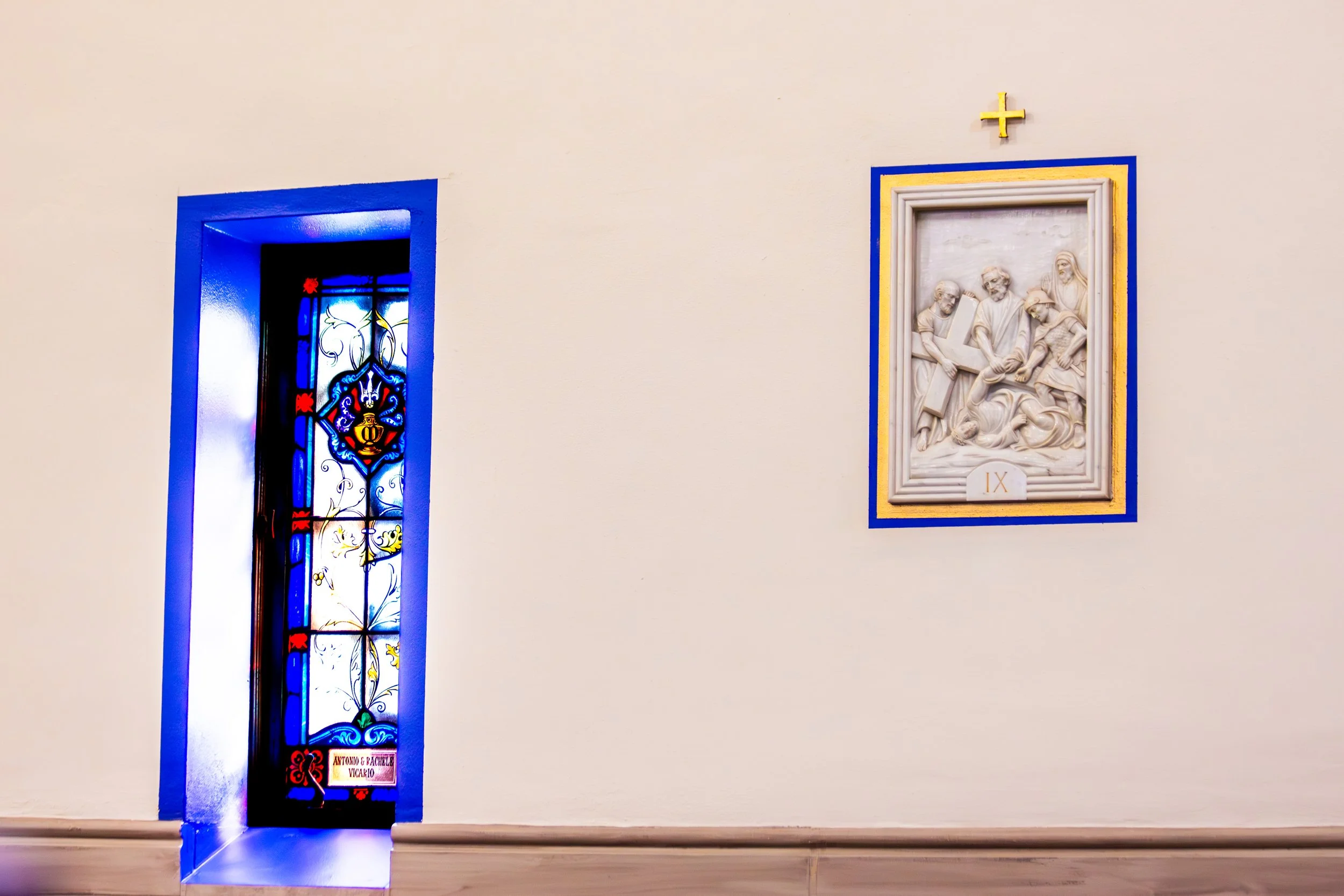 Interior of a church with a stained glass window on the left and a white relief sculpture on the right, which depicts a biblical scene with four figures. The relief is framed with blue and yellow borders, and there is a gold cross above it.