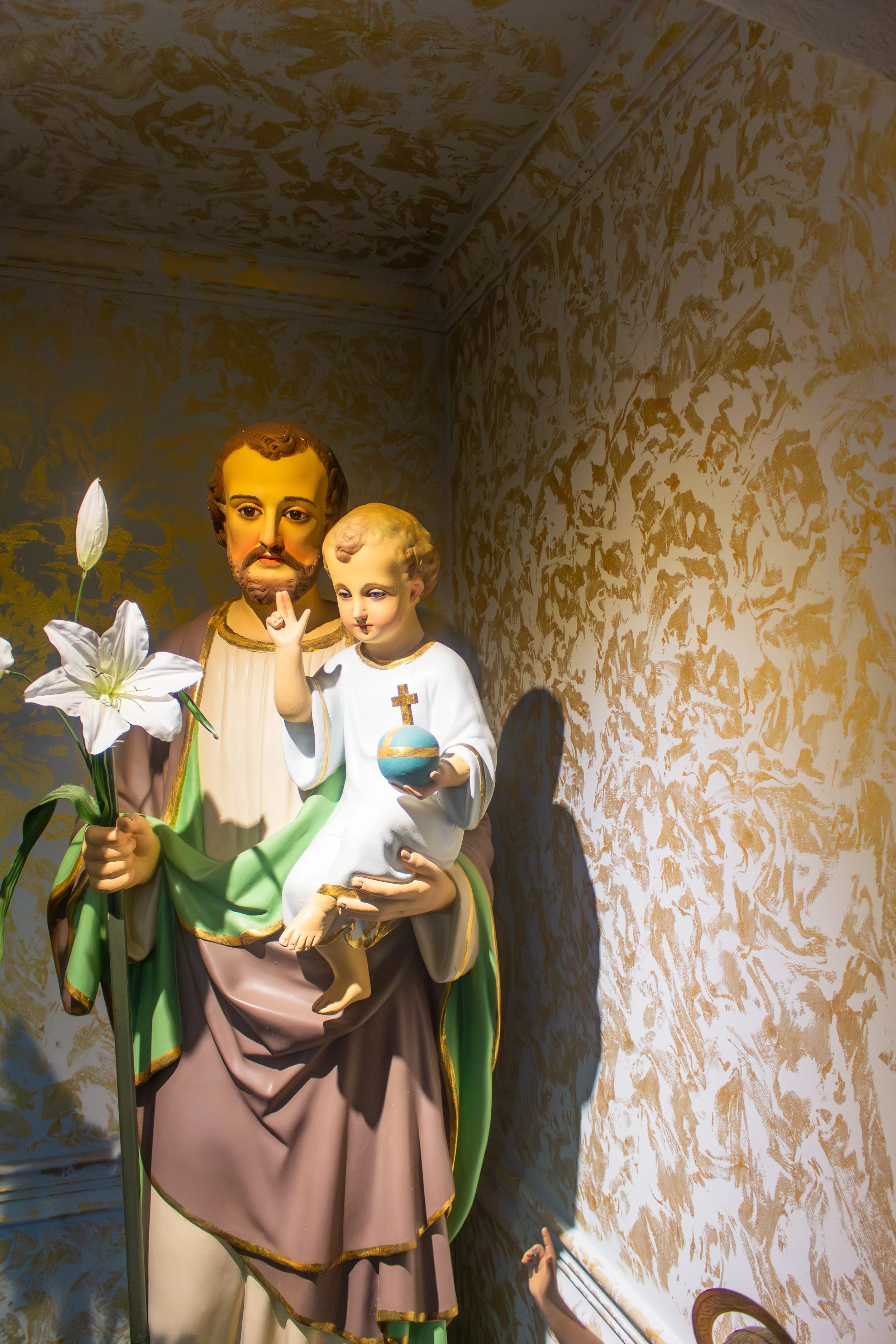 Statue of Saint Joseph holding baby Jesus, flowers, and a gold medallion, against a gold and cream textured background.