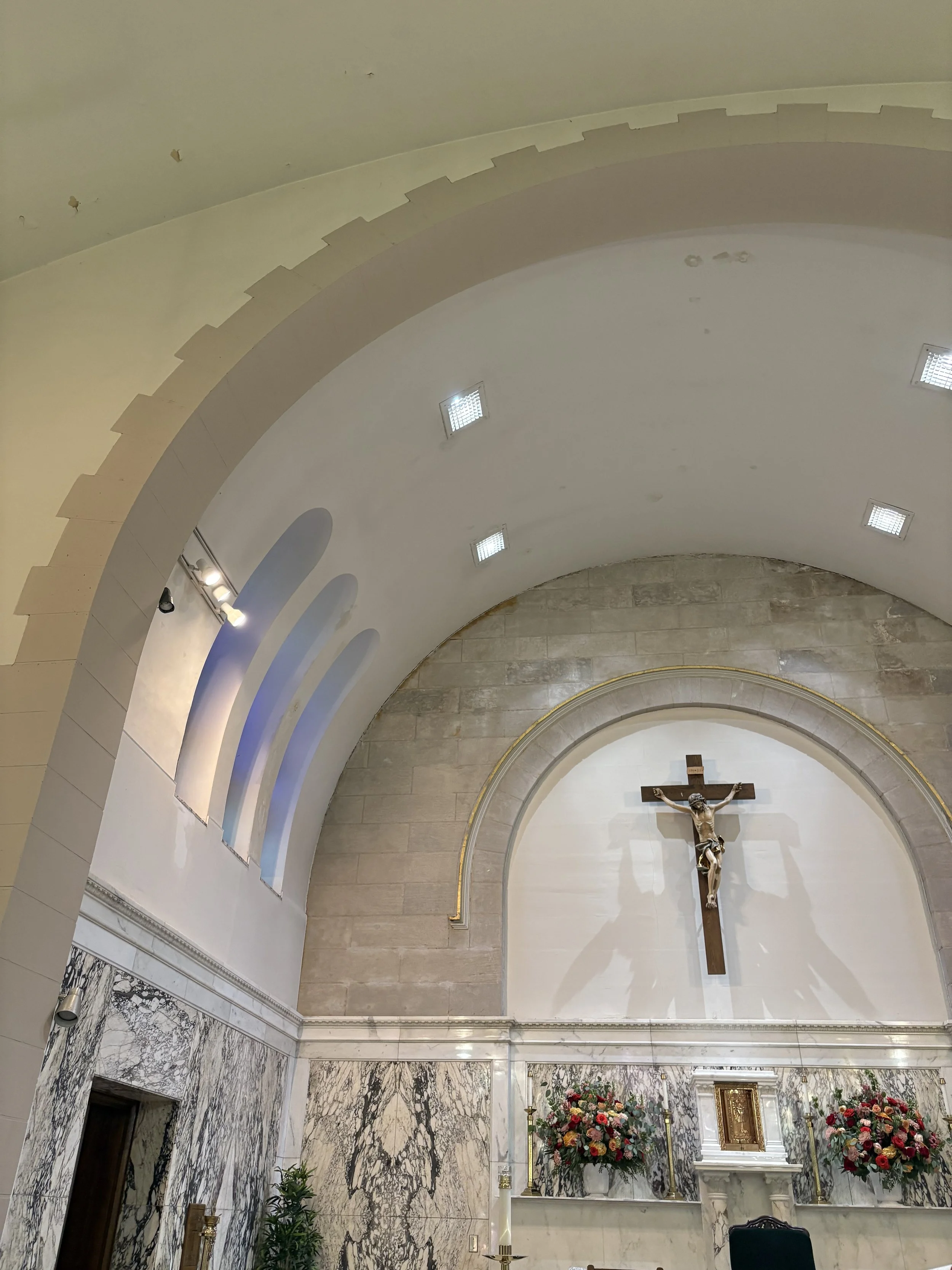 Interior view of a church or cathedral with a high arched ceiling, religious crucifix, floral arrangements, and marble walls.