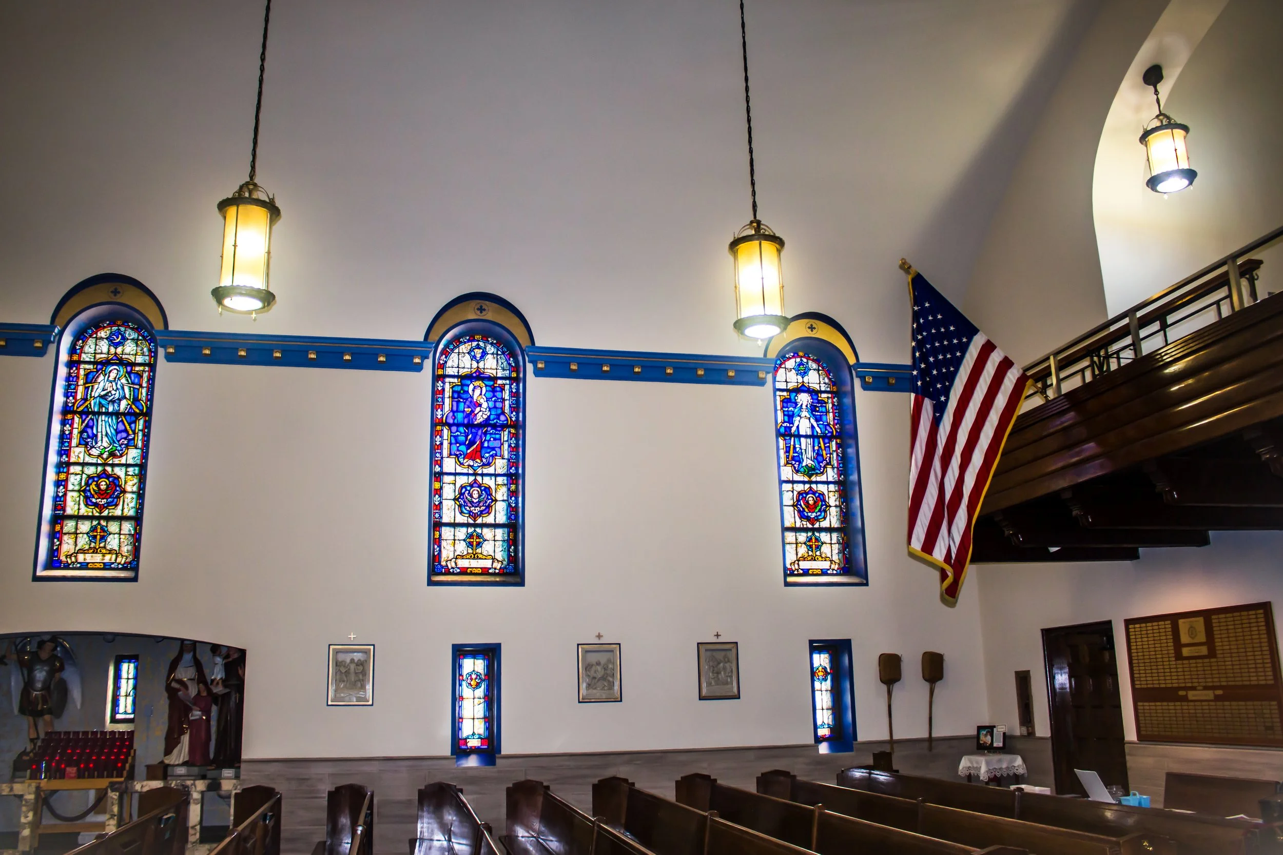 Interior of a church with stained glass windows, hanging pendant lights, an American flag, and pews.