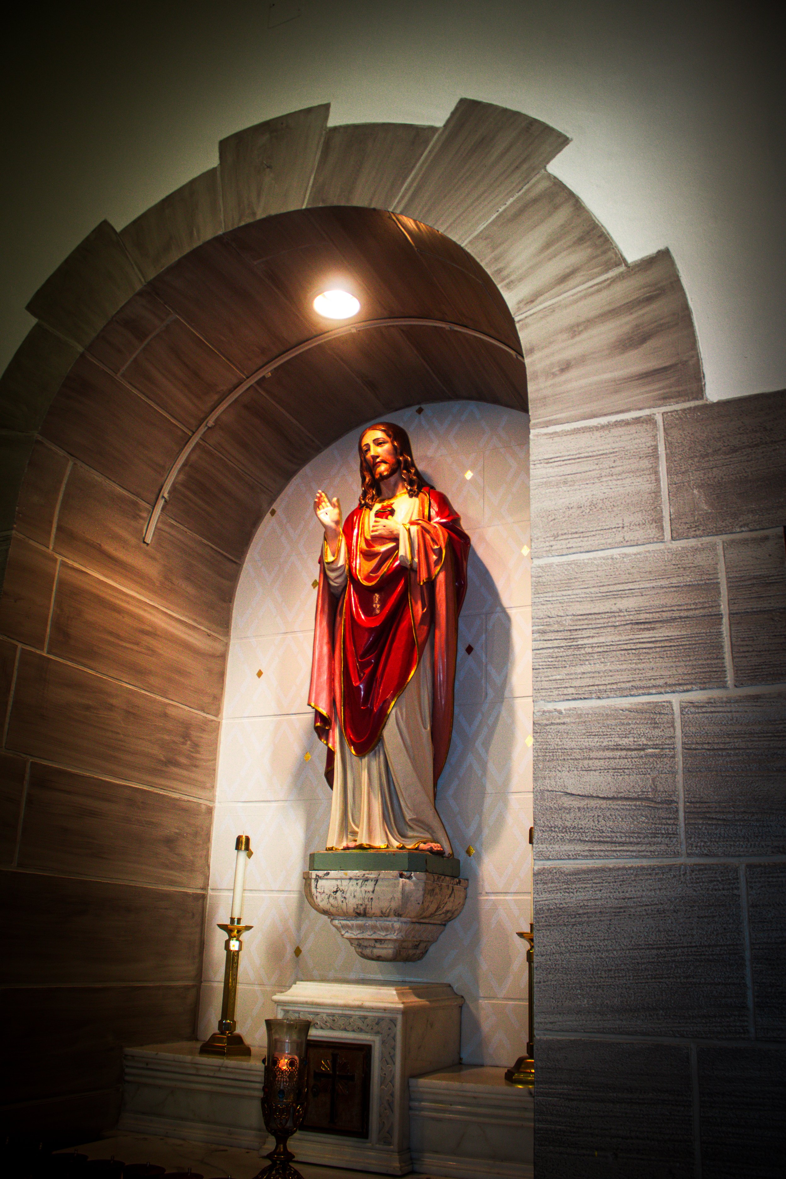 A religious statue of Jesus Christ with a red robe, standing on a pedestal in a church alcove, flanked by two candles.