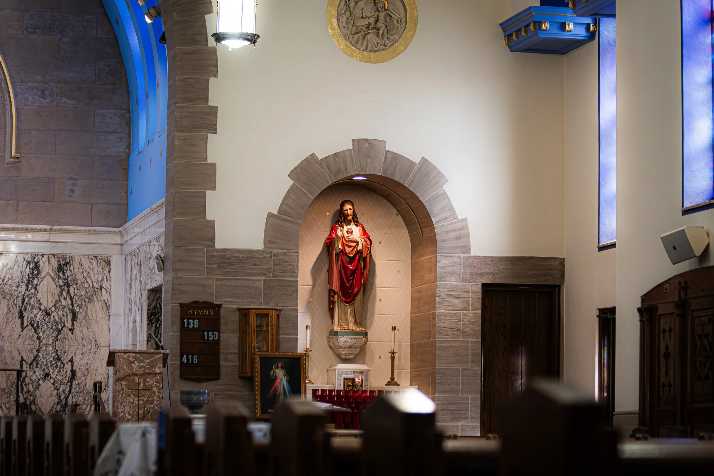 Inside a church, a religious statue of Jesus Christ holding a sacred heart, set within a semi-circular stone arch, with wooden pews and altar decorations including candles and religious images.