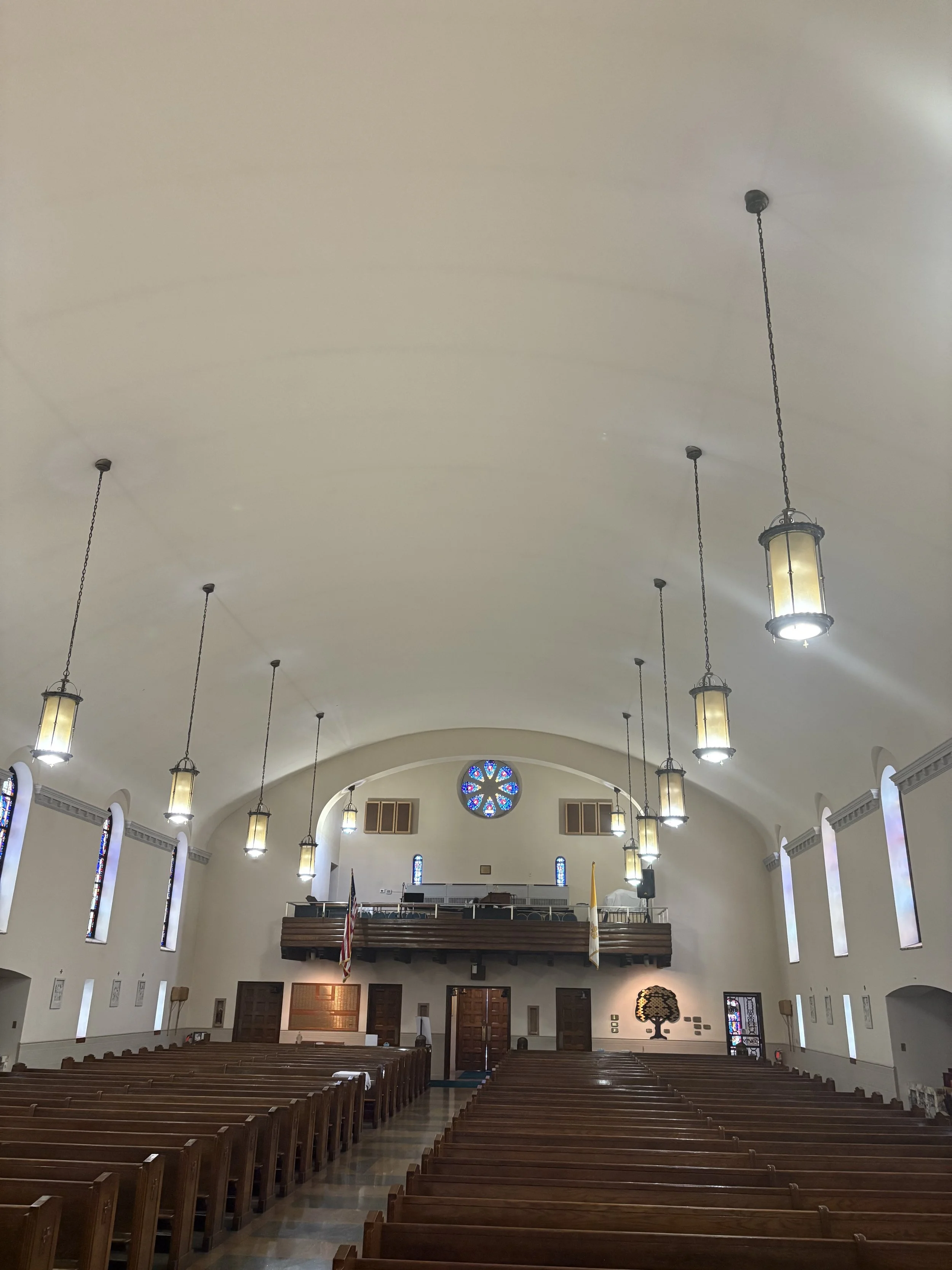 Interior of a church with wooden pews, stained glass windows, hanging lantern lights, and a high arched ceiling.
