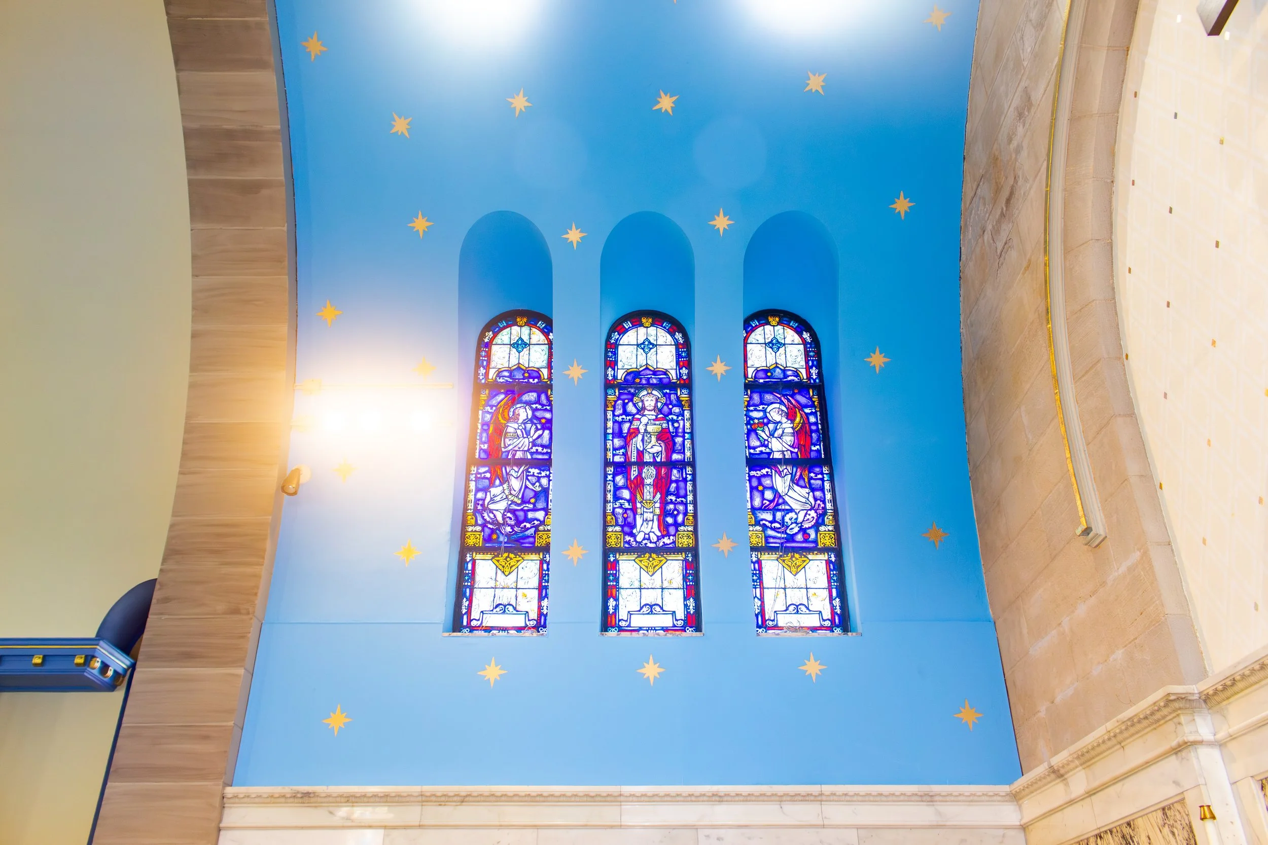 Interior of a church with three stained glass windows depicting religious figures, a bright blue wall with gold star decorations, and a section of marble flooring.