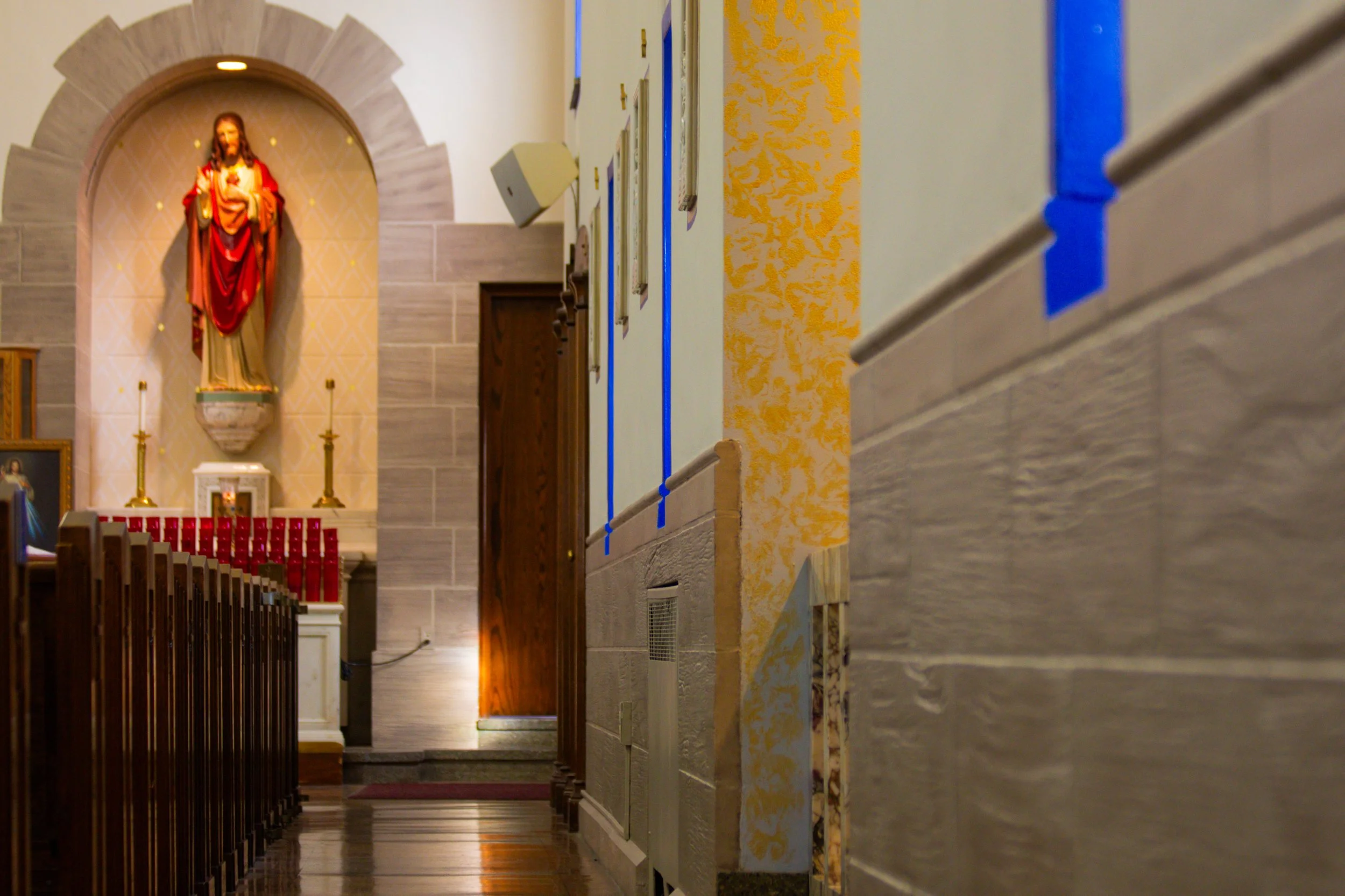 Interior of a church with a statue of Jesus Christ in a niche, wooden pews, and yellow textured wall on the right.