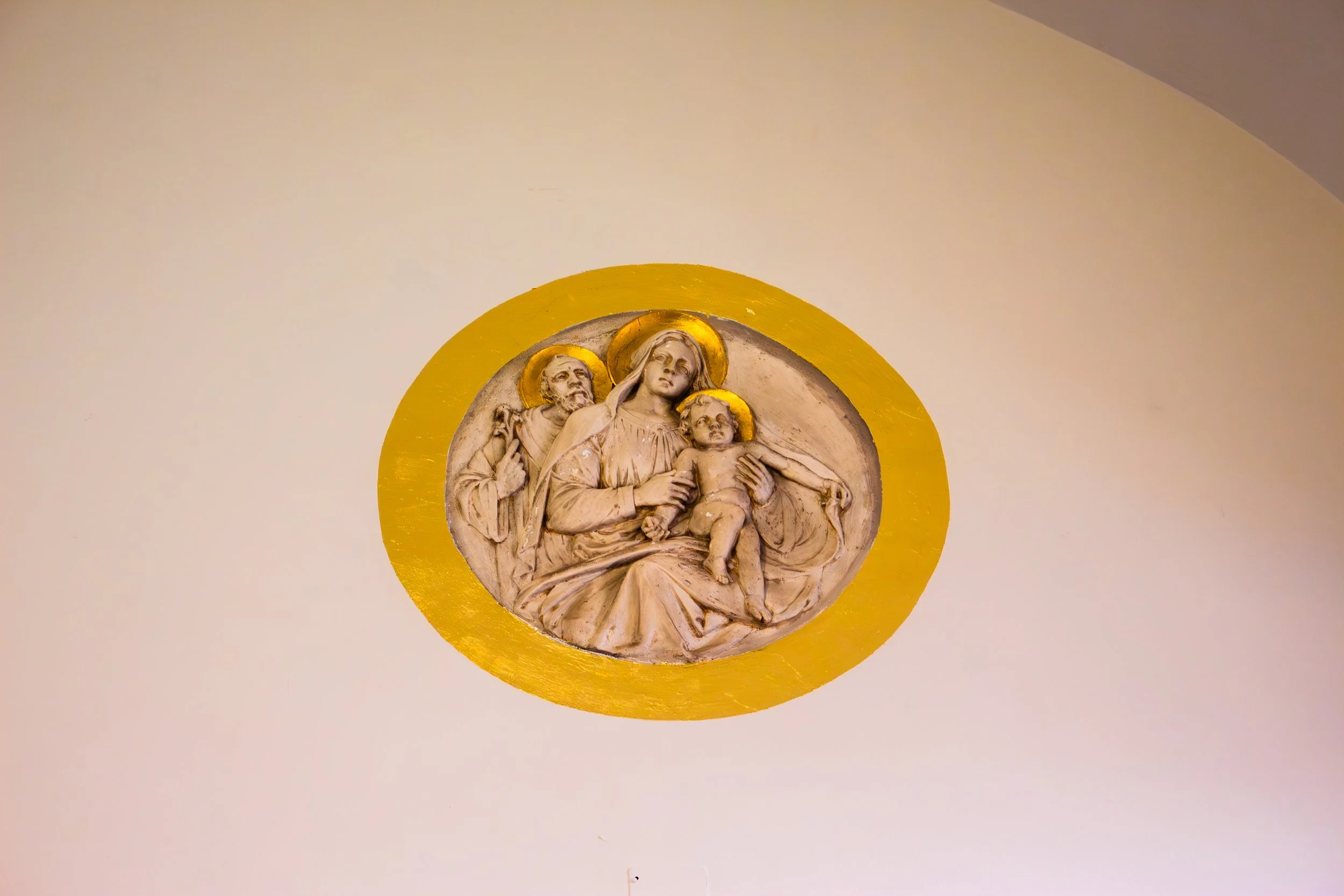 Religious bas-relief sculpture of the Virgin Mary with Jesus and Saint Joseph, framed with a yellow circular background, mounted on a plain white wall.