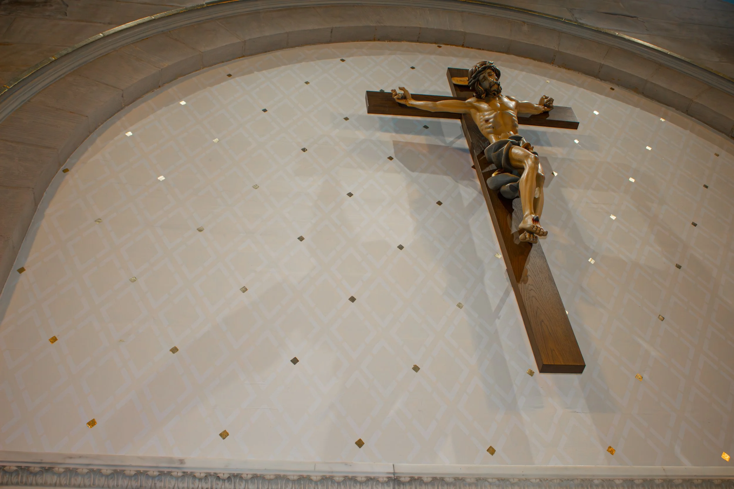 A wooden crucifix with Jesus Christ on it, mounted on an arched white wall with small gold and black decorative tiles, inside a church or cathedral.
