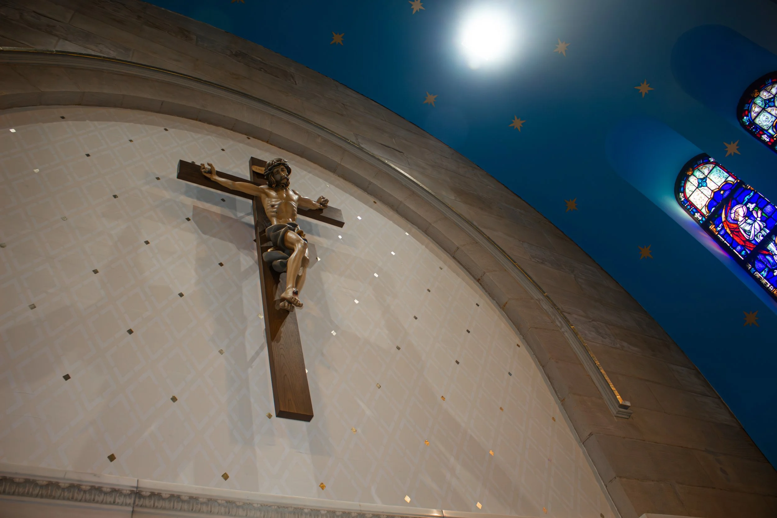 Interior of a church with a wooden crucifix of Jesus Christ on a wall, stained glass windows, and decorated blue ceiling with gold stars.