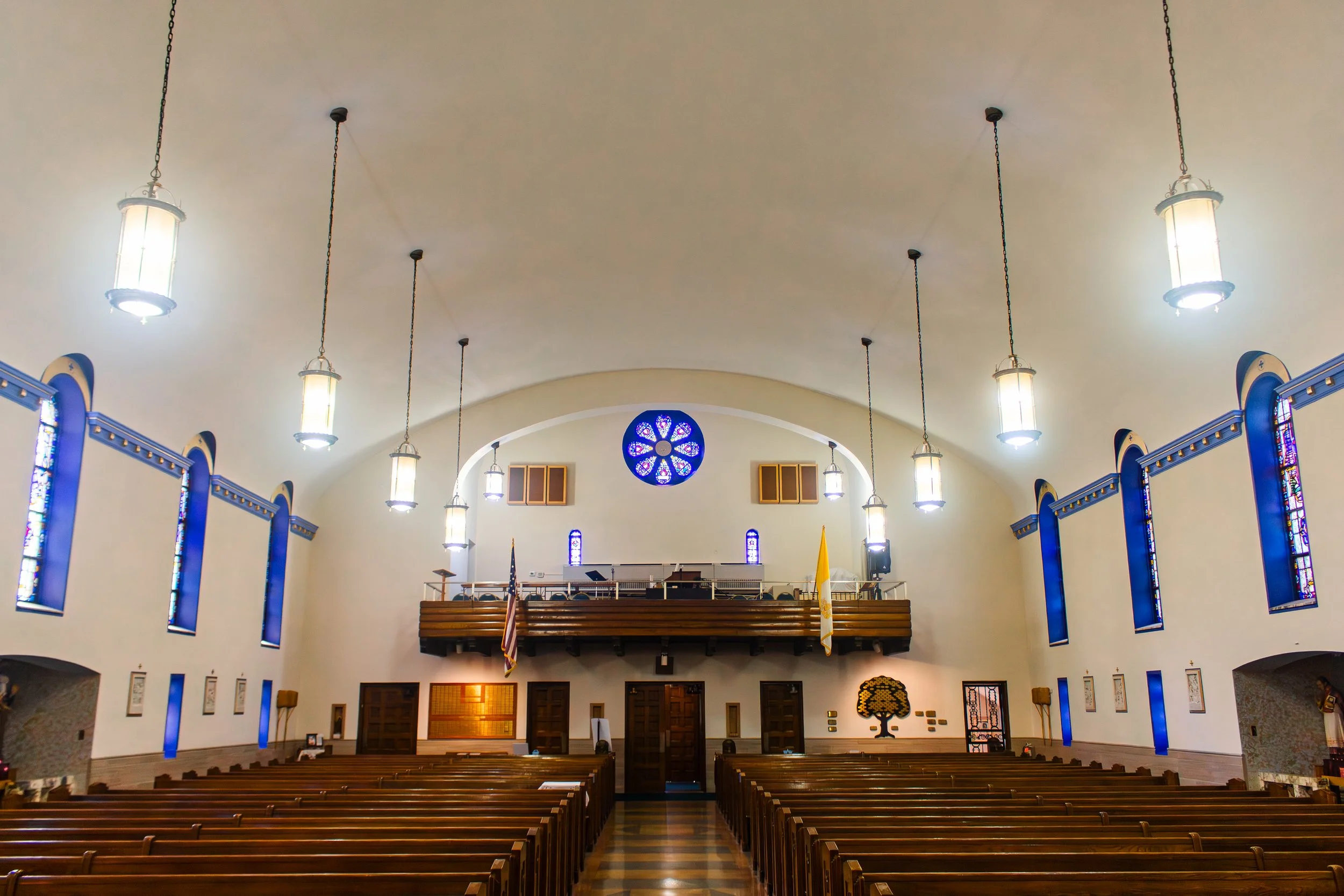 Interior of a church with wooden pews and blue stained glass windows. Hanging lights are suspended from the ceiling. There is a circular stained glass window at the front, above the altar area.