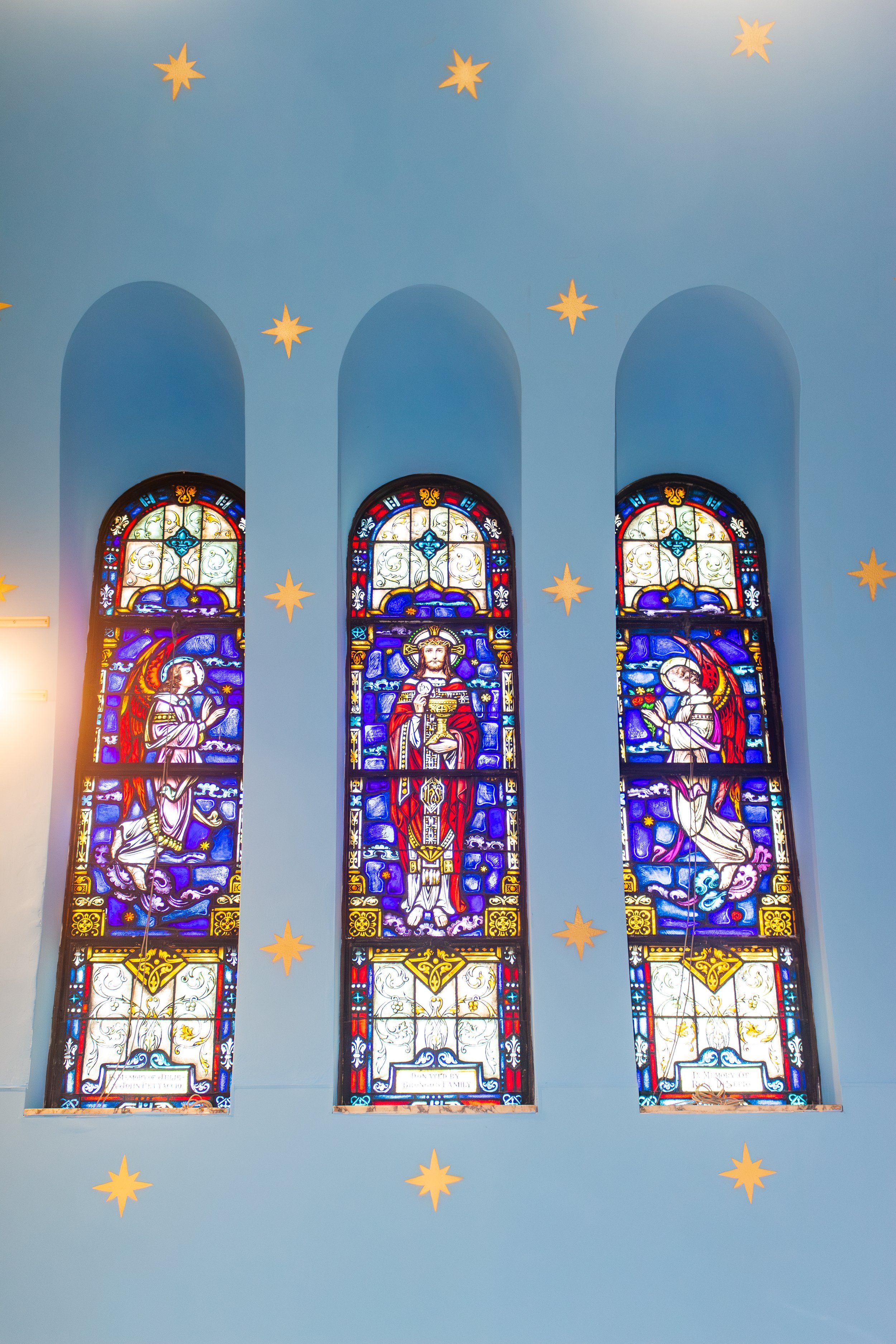 Interior of a church with three tall stained glass windows depicting religious figures, mounted on a light blue wall with star-shaped decorations.