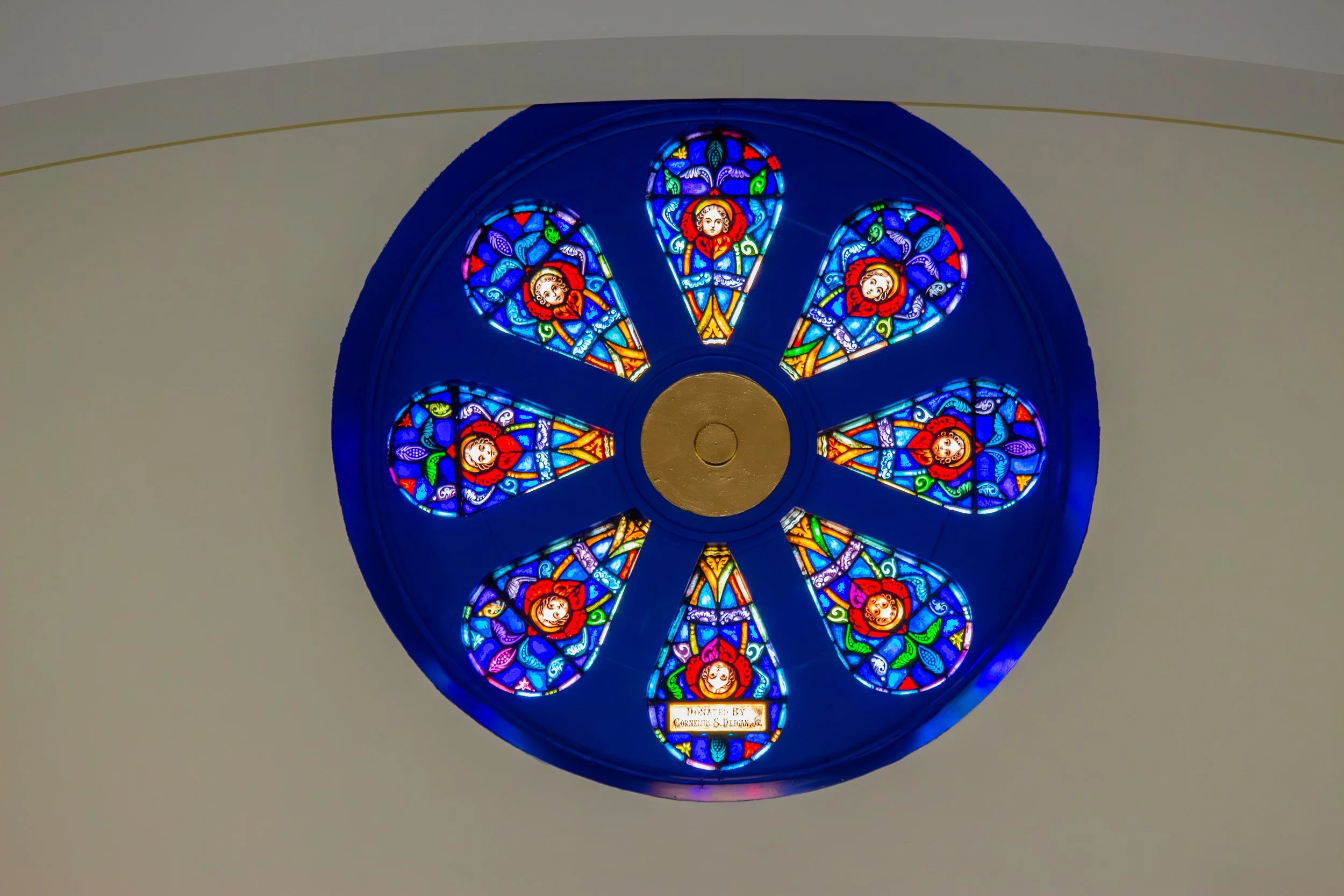 Colorful circular stained glass window featuring floral and face motifs, with a plaque at the bottom of the window.