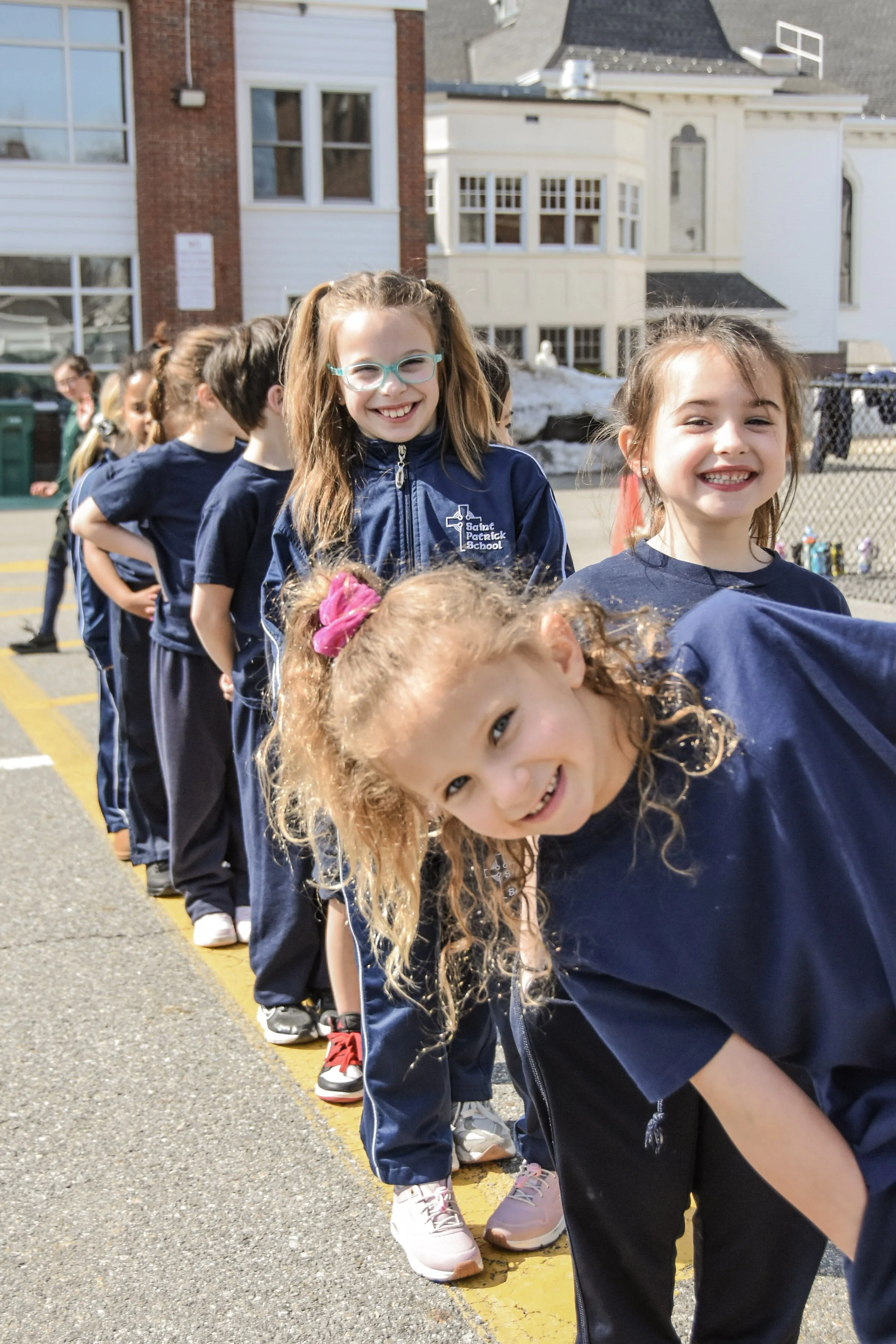 Smile and happy children in line outdoors on a schoolyard, dressed in navy blue uniforms, with buildings and snow in the background.