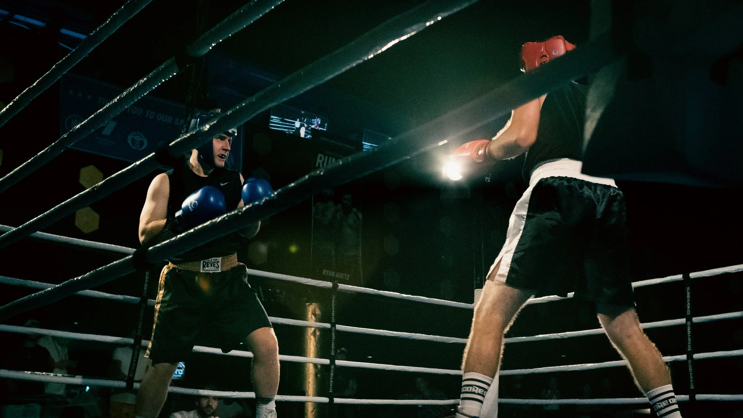 A boxing match featuring a woman and a man inside a dark boxing ring with ropes, with the woman wearing blue gloves and shorts, and the man wearing red gloves and black shorts. They are both in fighting stances, with the woman's expression intense and focused.