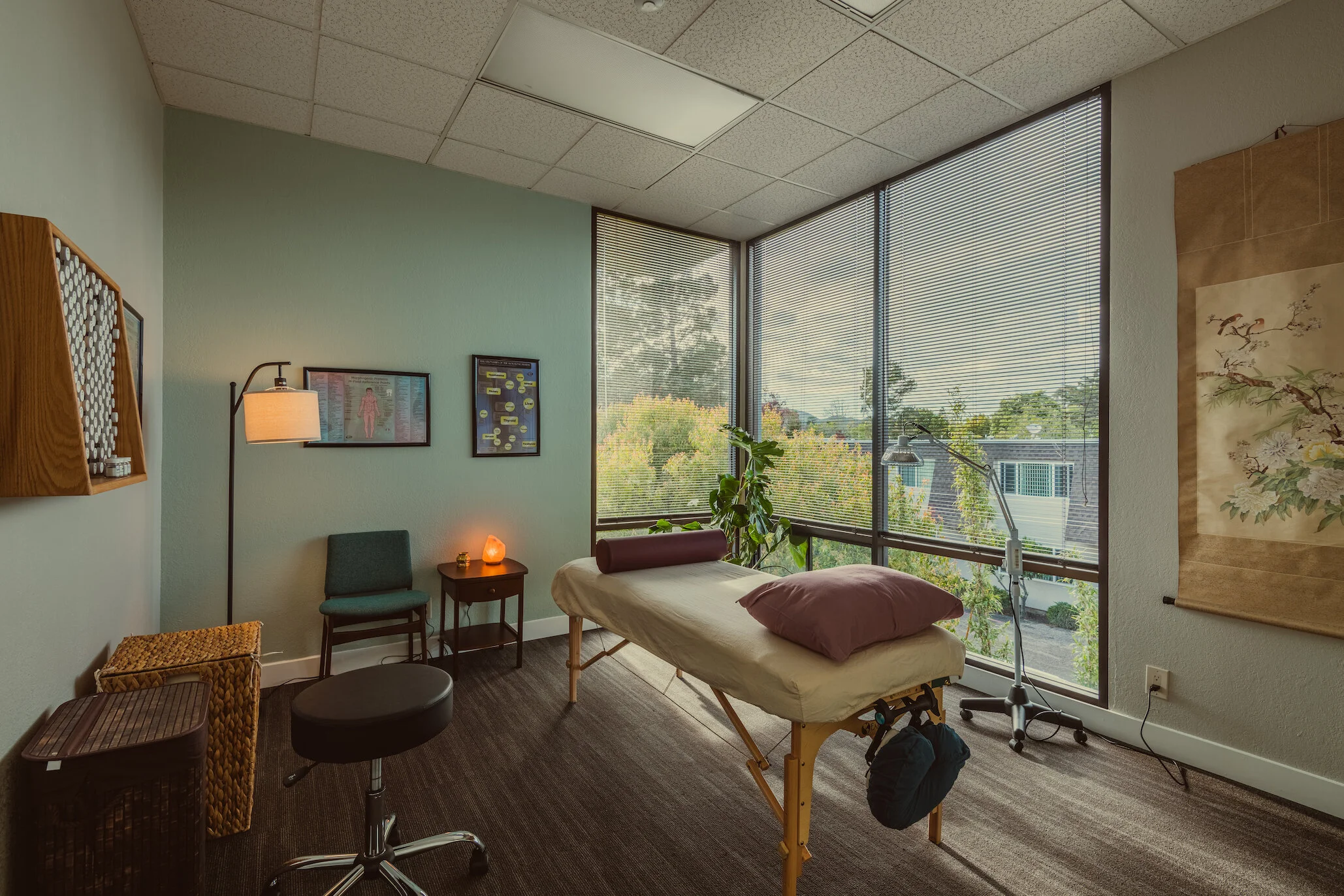 A massage therapy room with a massage table near large windows with blinds, a pillow, a bolster, and a plant. The room has framed artwork and a salt lamp on a small side table, with a floor lamp, a chair, a small stool, and decorative baskets.