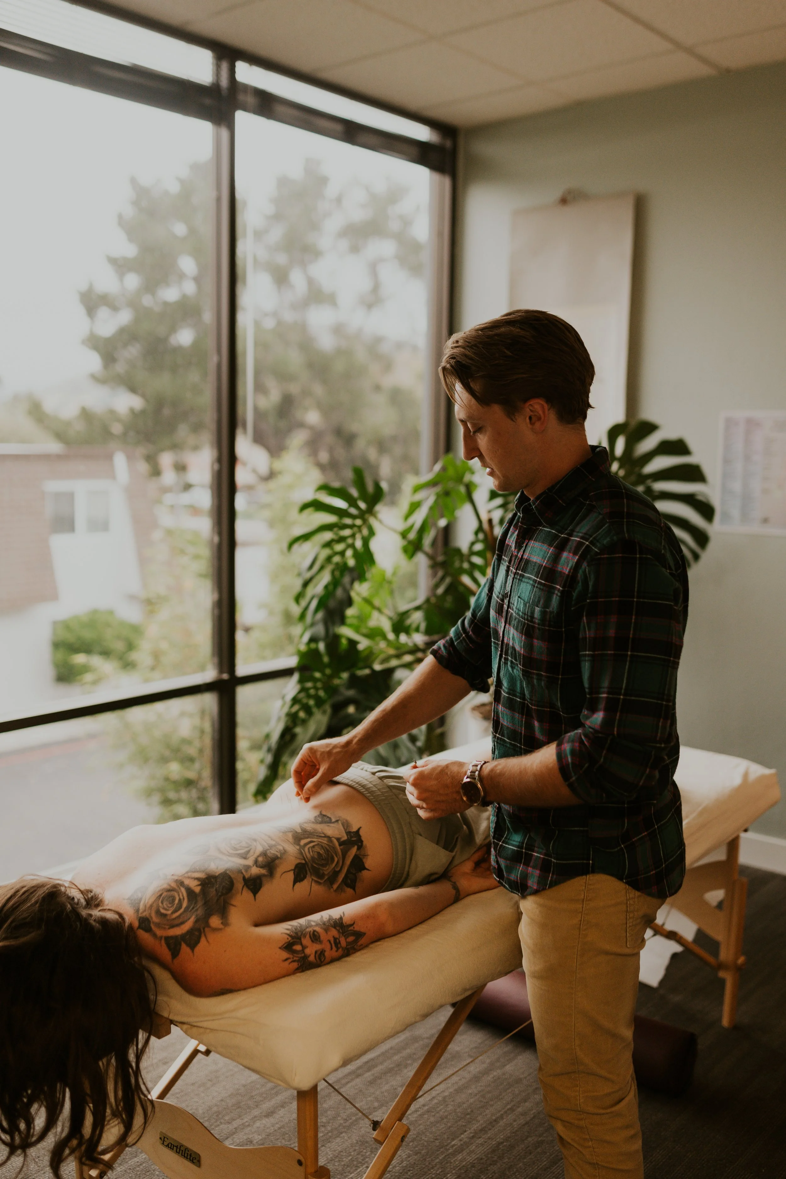 A man giving a massage to a woman lying face down on a massage table in a room with large windows and plants.