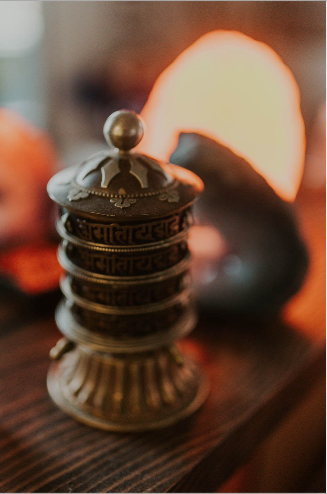 Stack of six brass prayer wheels with a dome-shaped top and a small brass ball, with blurred pumpkins and a glowing orange pumpkin slice in the background.