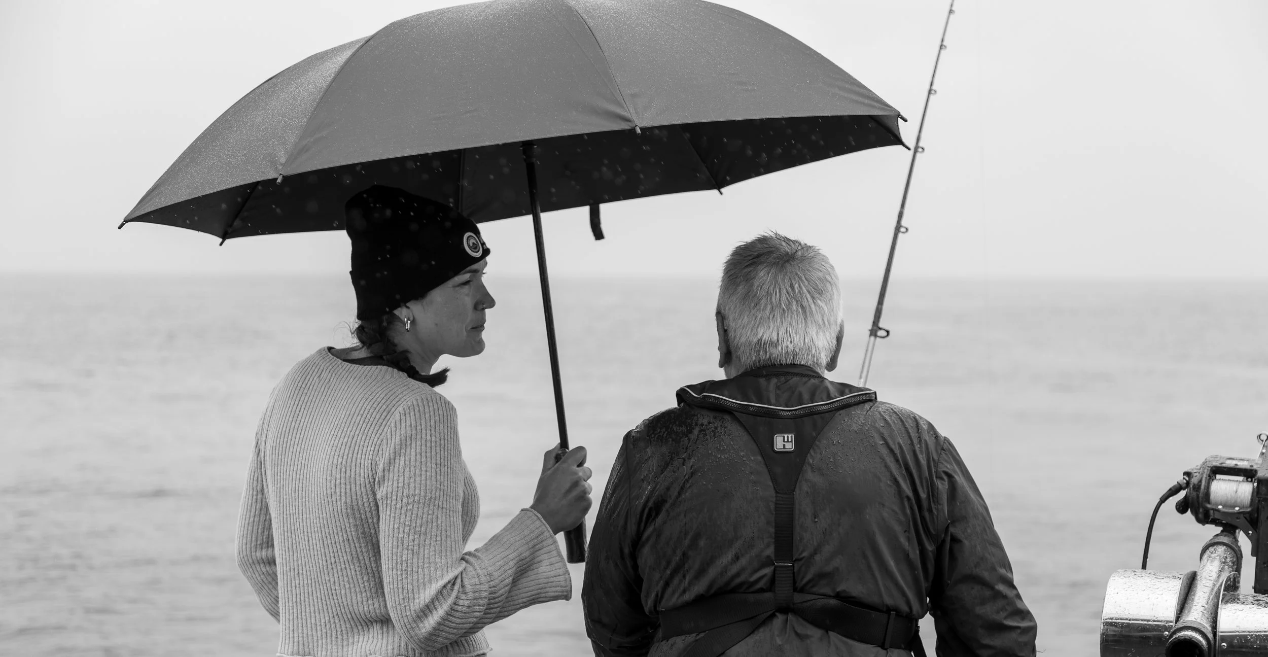 Black and white photo of a woman and man standing on a boat, sharing an umbrella, with the woman speaking to the man.