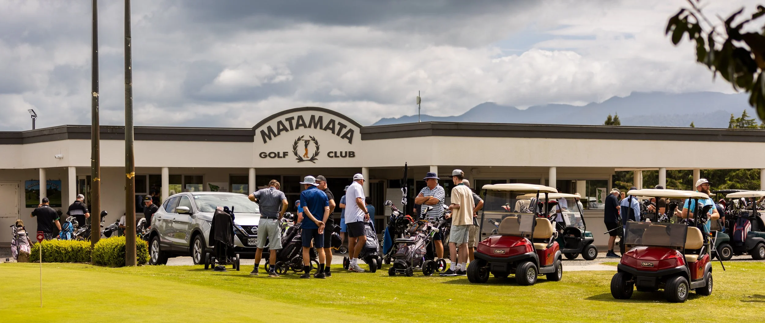 A group of people gathered outside the Matamata Golf Club building, with golf carts and golf bags visible, on a partly cloudy day with mountains in the background.
