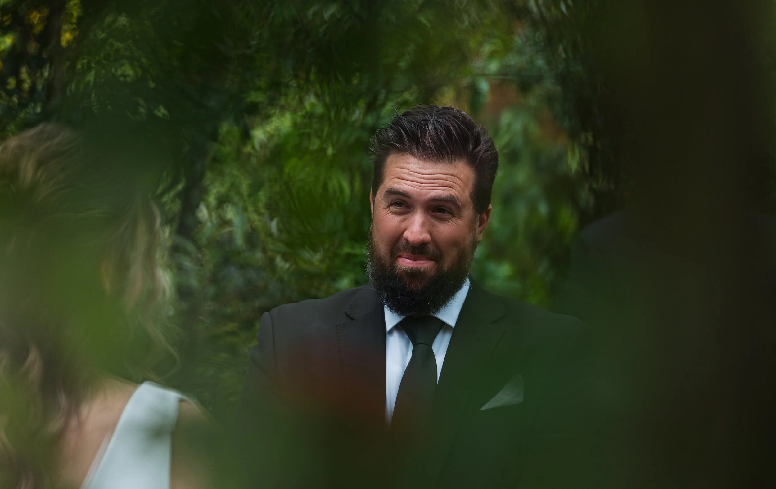 Man with dark hair and beard wearing a black suit and tie, talking to a woman outdoors with blurred green foliage around.