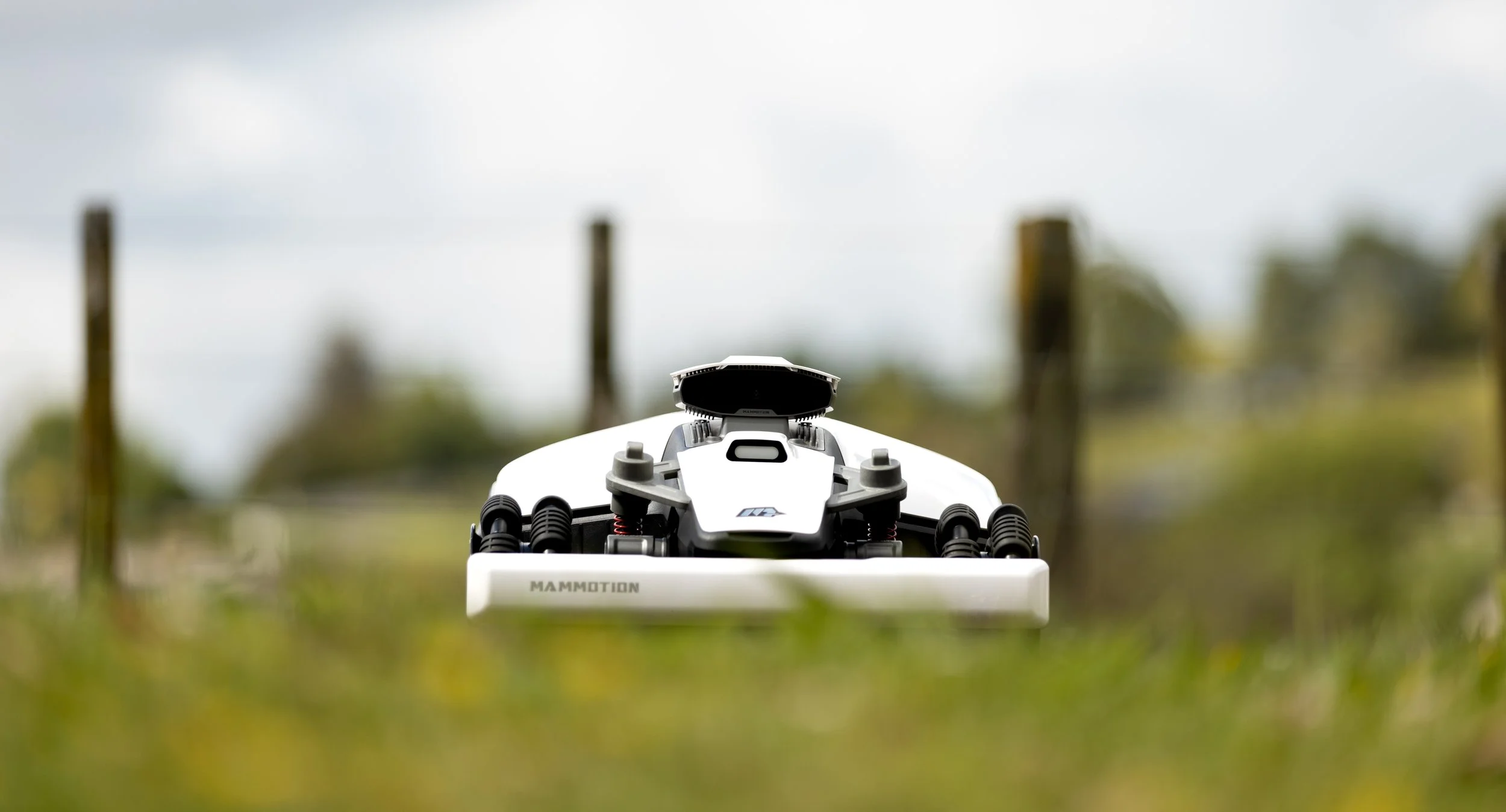 Close-up of a white drone on grass with blurred background and wooden posts.