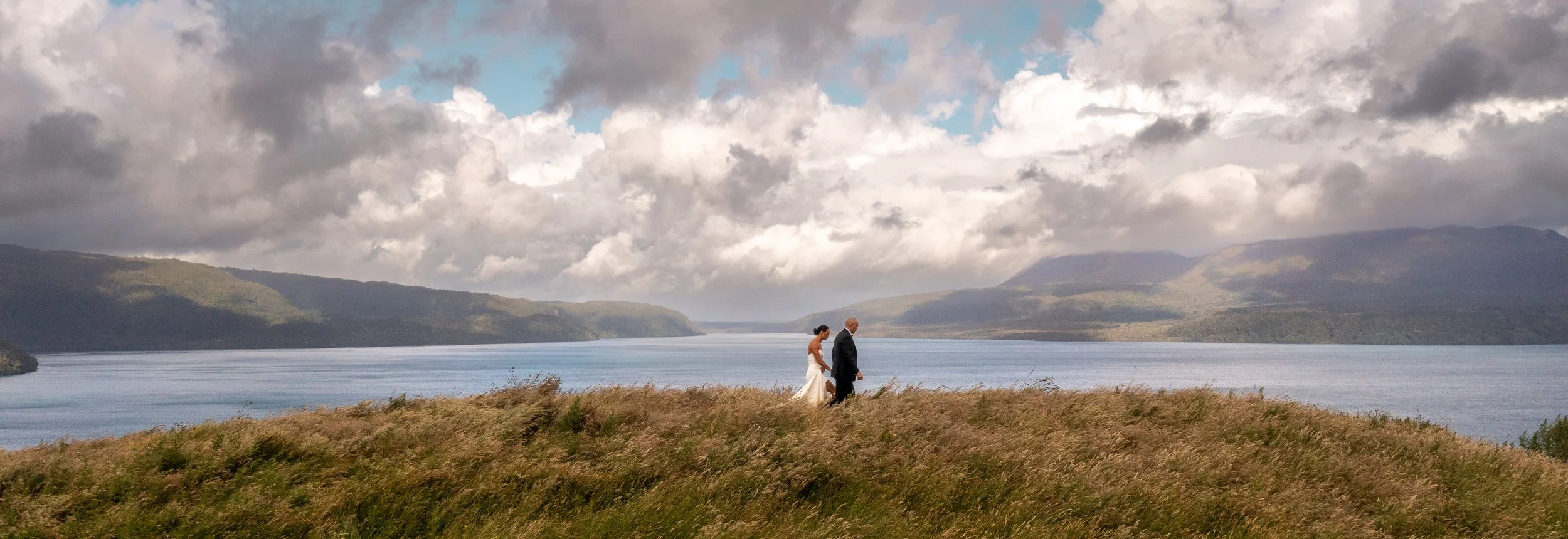 A bride and groom walking together on a grassy hill by a large lake or river, with mountains and a cloudy sky in the background. Blackbarn wedding venue