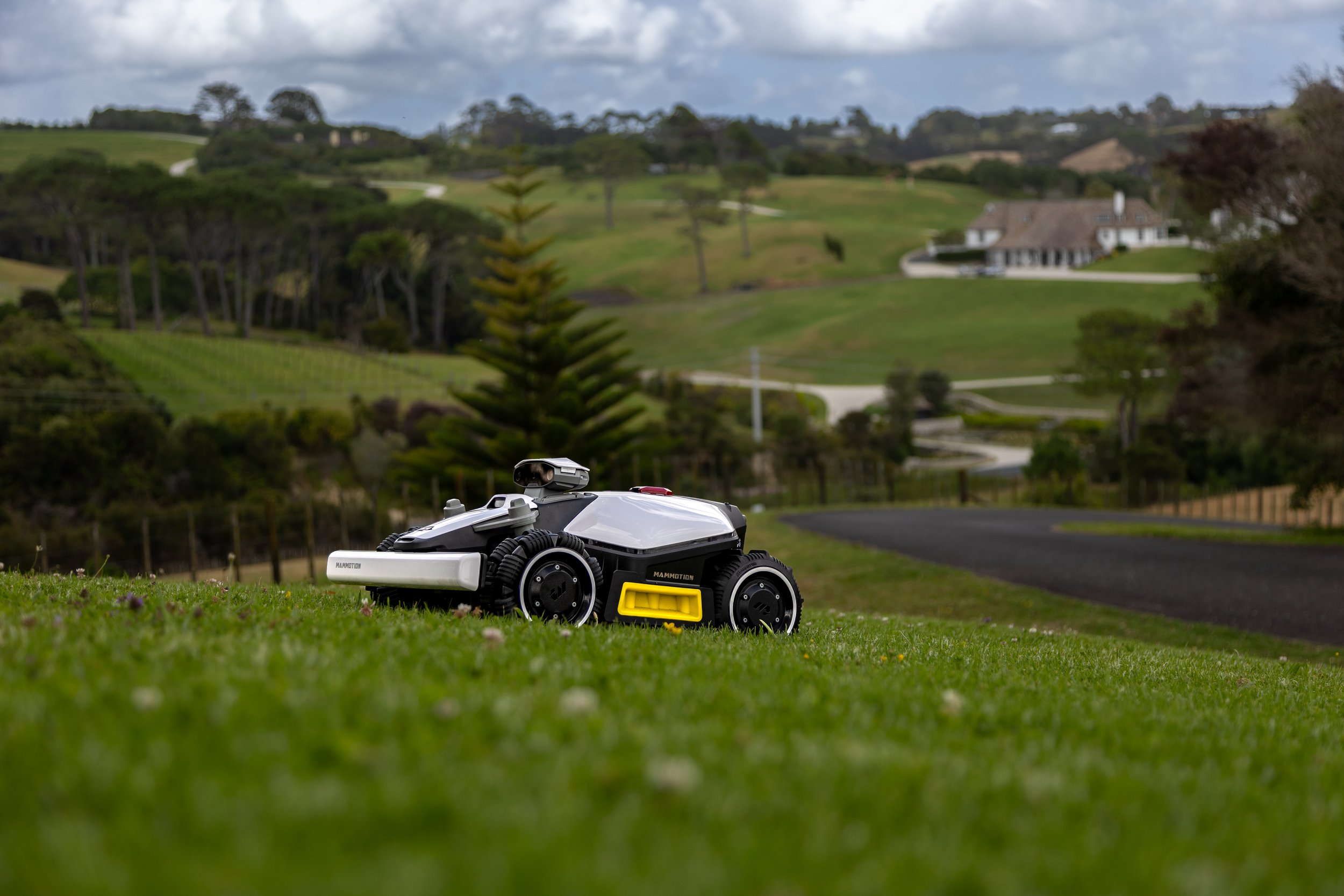 A robotic lawn mower on a grassy lawn with a scenic countryside with trees, houses, and rolling hills in the background.