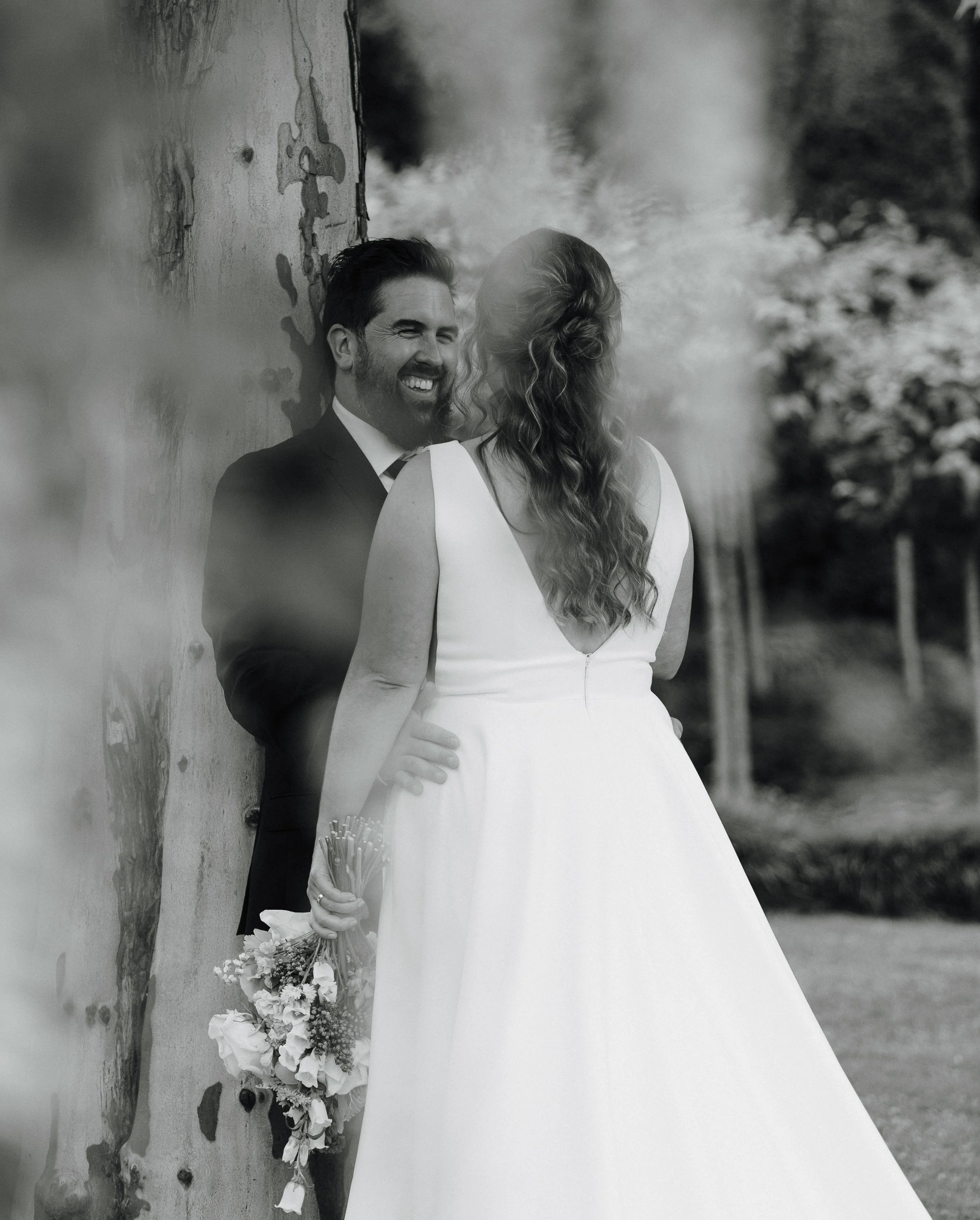 A black and white photo of a bride and groom outdoors. The bride faces away from the camera, holding a bouquet, and the groom leans against a tree, smiling at her. Tree church wedding venue