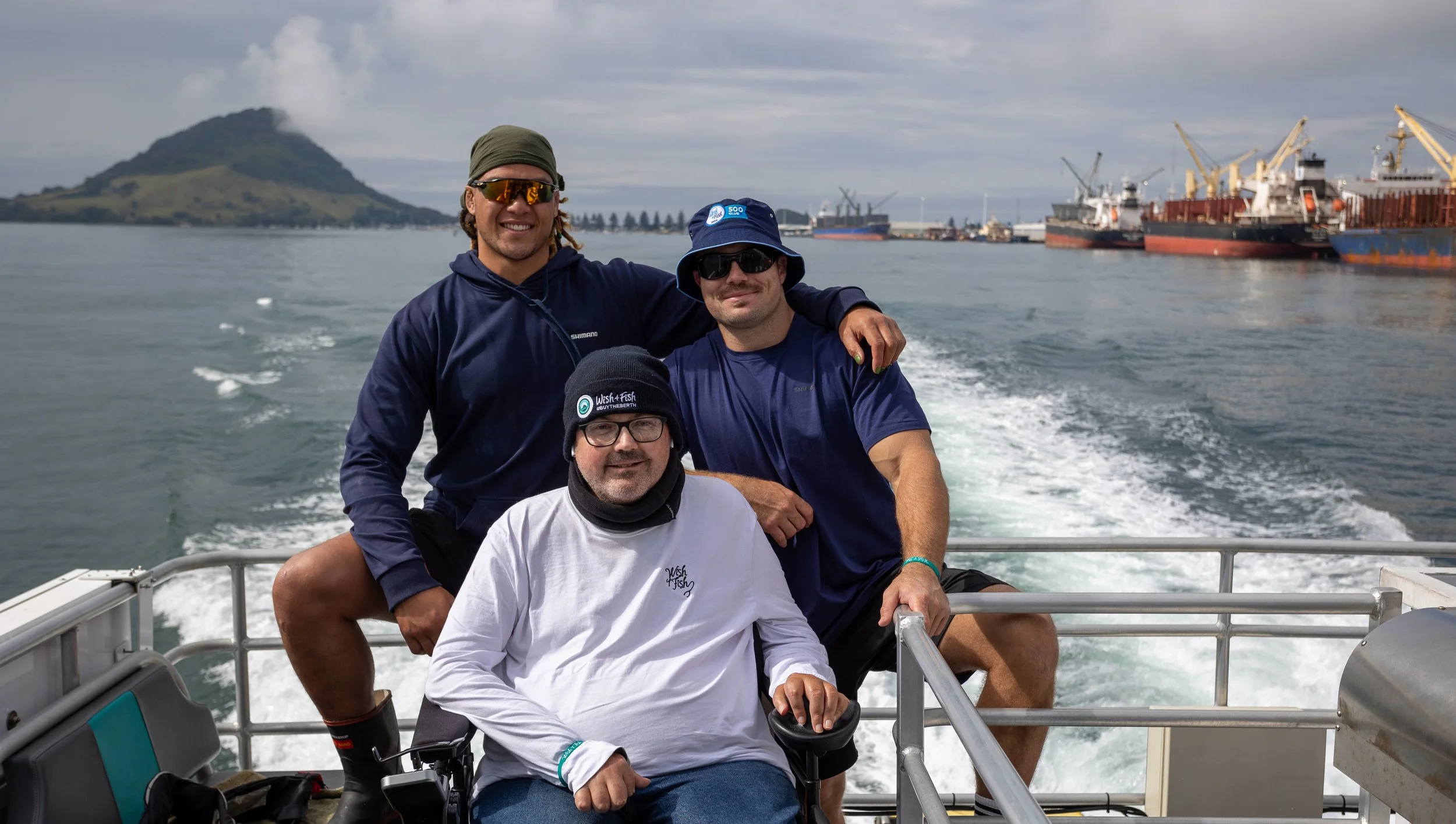 Three men on a boat with a harbor, ships, and a mountain in the background.